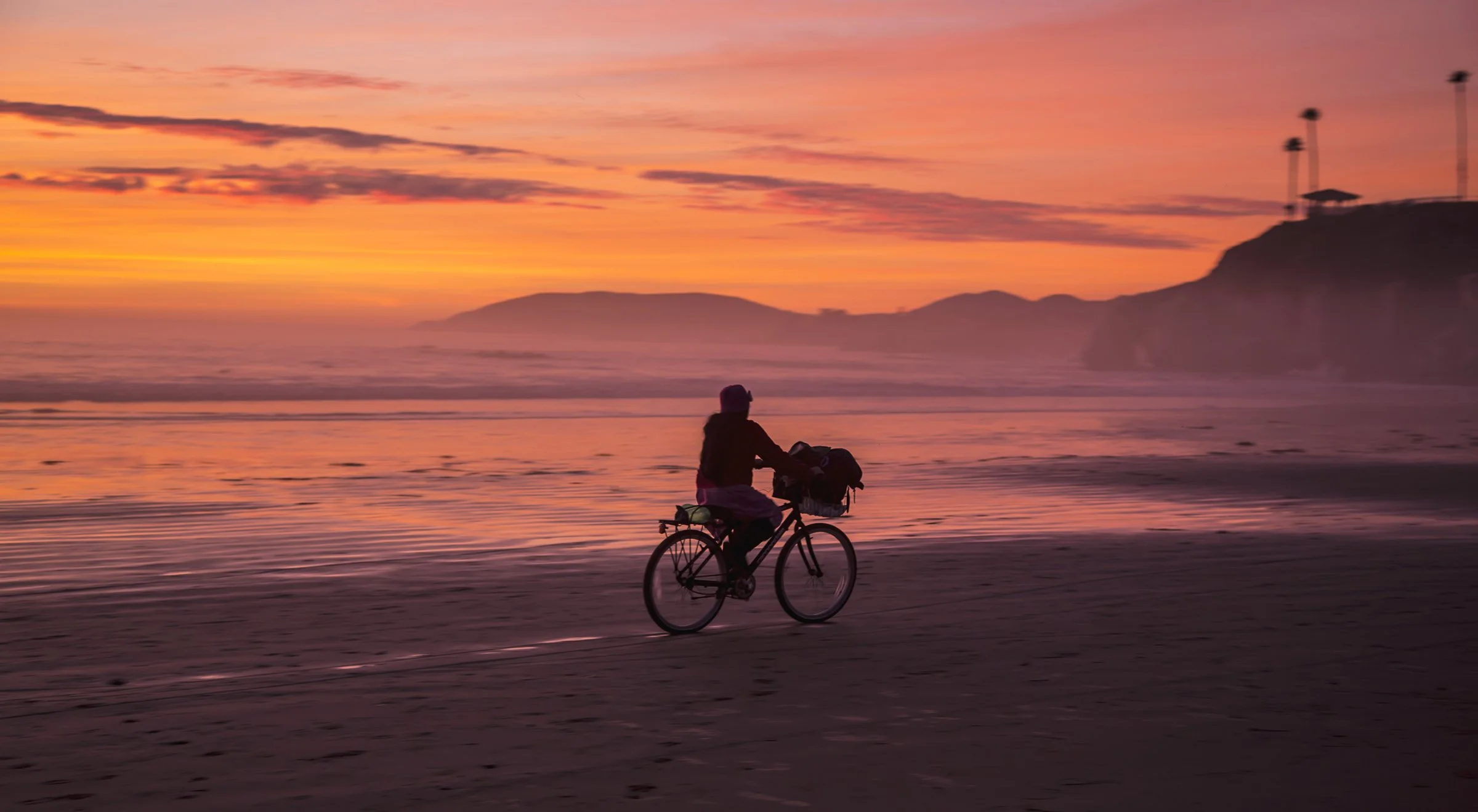 Person biking along the beach at sunset with colorful sky, ocean, and distant cliffs.