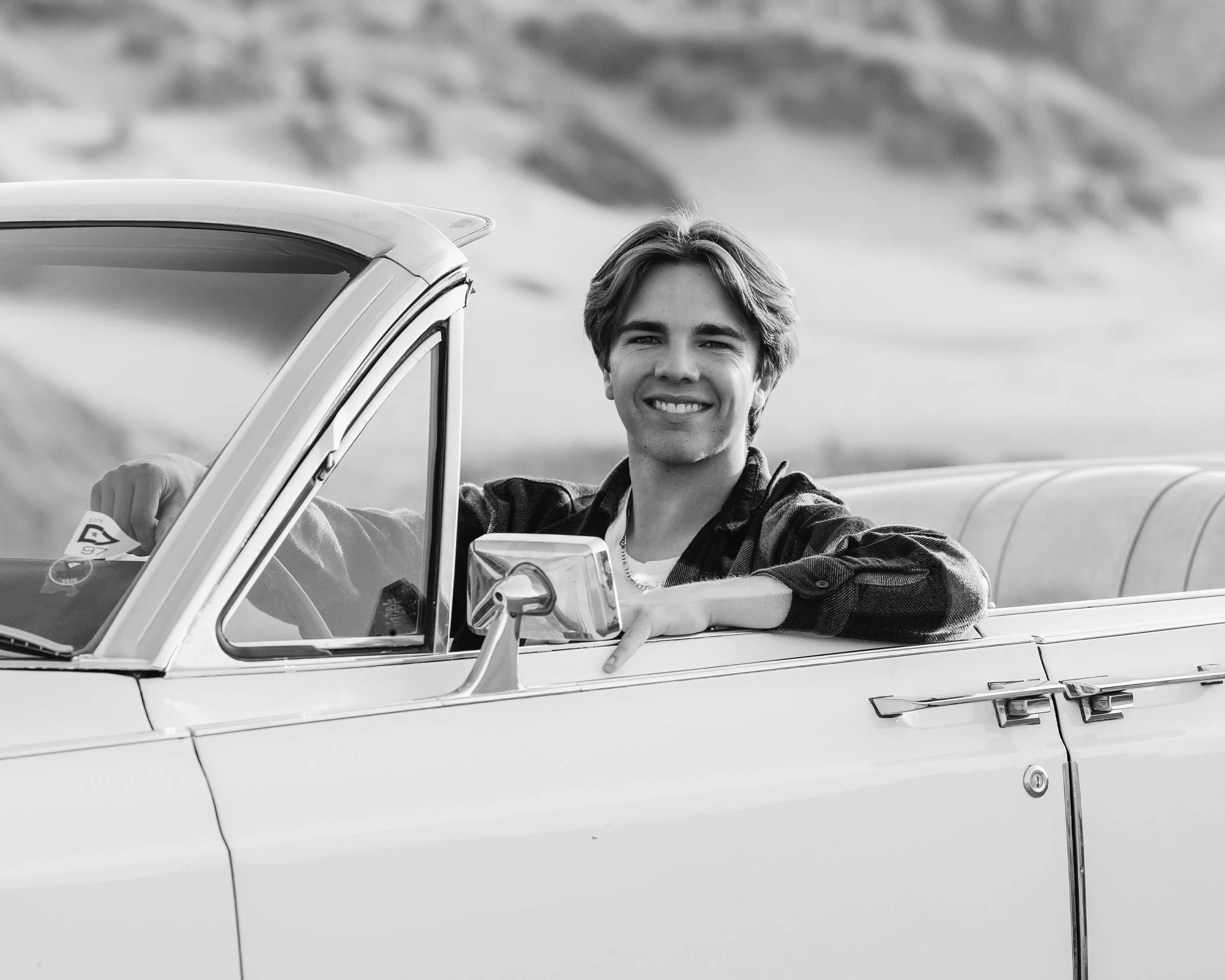 A young man with light skin, smiling, leaning out of a vintage car window with a landscape of mountains or hills in the background.