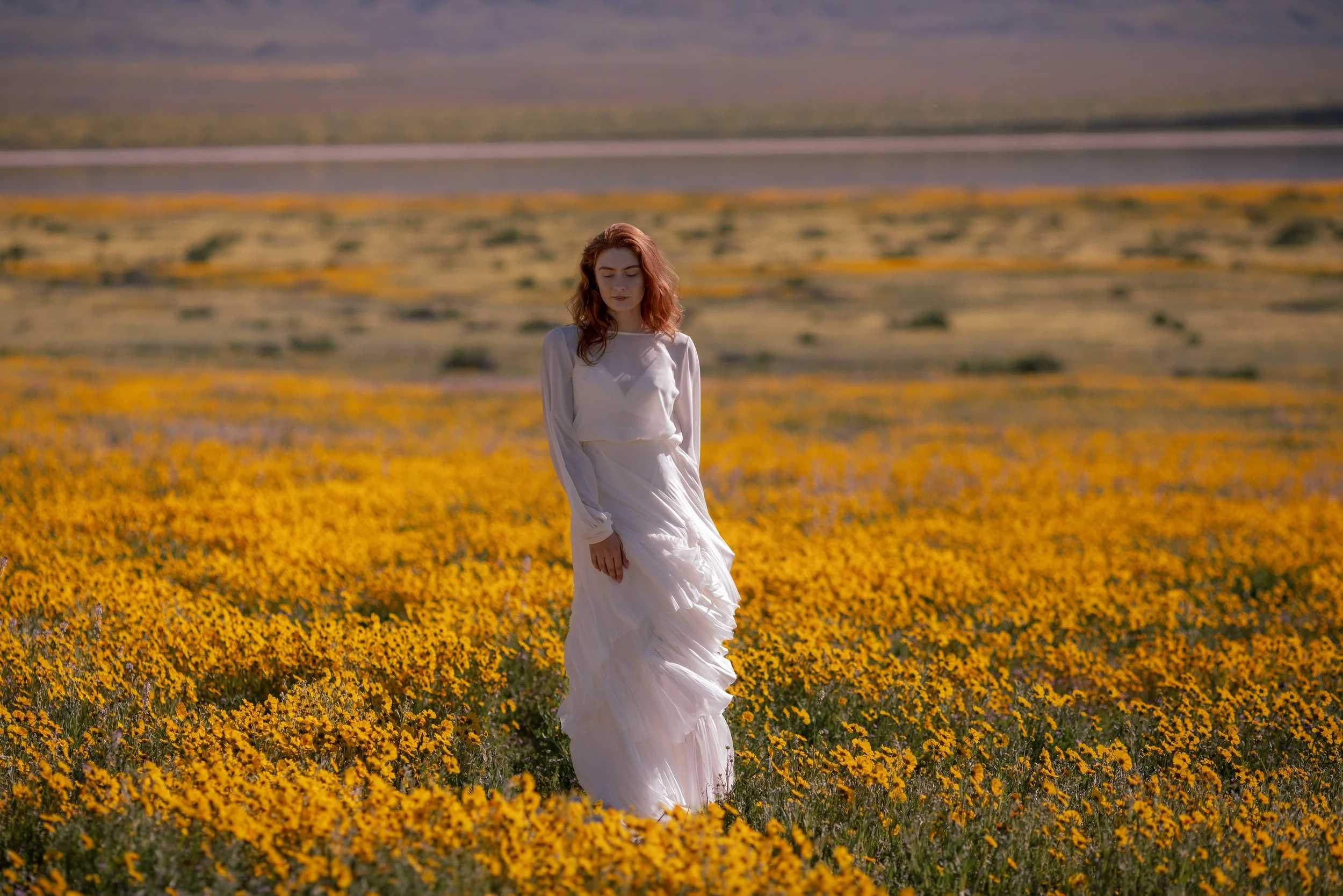 A woman in a white dress walking through a field of yellow and purple flowers, with mountains and a body of water in the background.