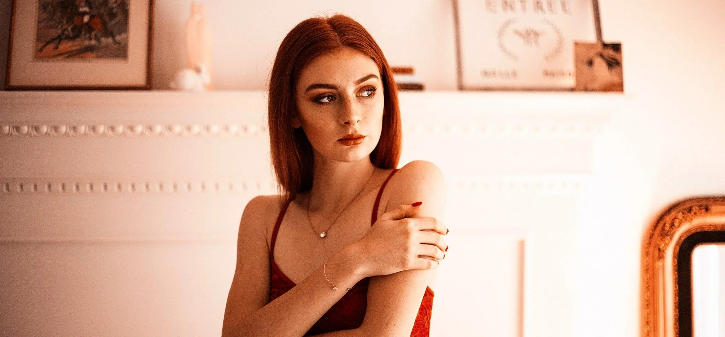 A young woman with red hair and makeup wearing a red dress and jewelry, standing indoors near a wall with framed artwork.