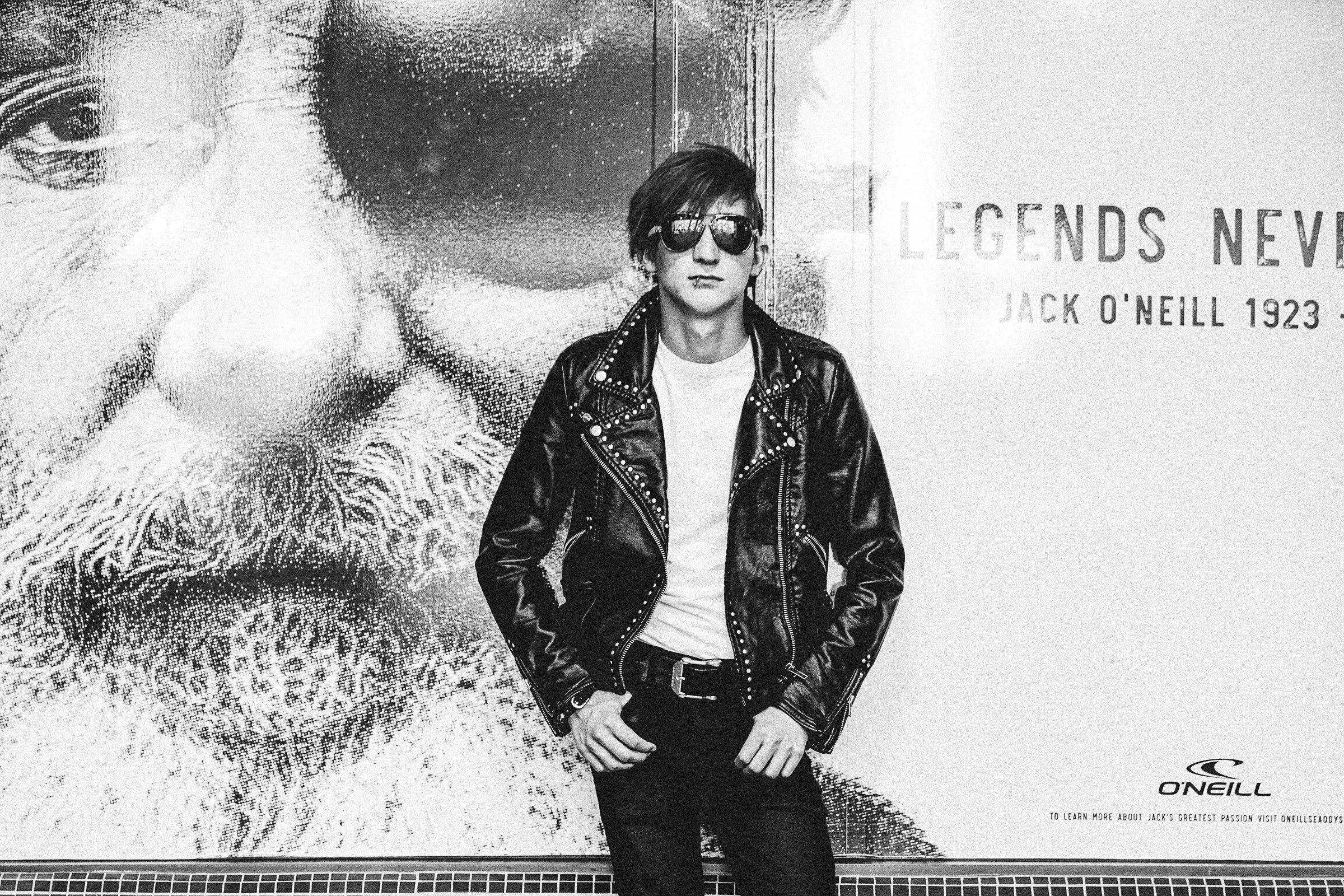 A young man with short hair, wearing dark sunglasses, a lip piercing, a white T-shirt, and a studded leather jacket, stands in front of a large black and white poster of Jack O'Neill, with the text 'LEGENDS NEVER' and 'JACK O'NEILL 1923.' The man has a neutral expression and is facing the camera.