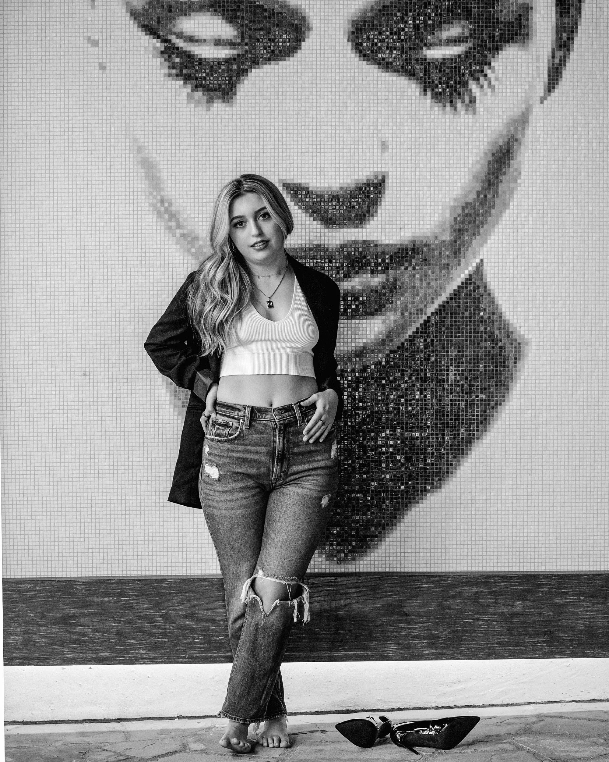 A woman standing in front of a mosaic wall with an abstract face design, wearing a white top, ripped jeans, and a black jacket, with shoes on the floor beside her.