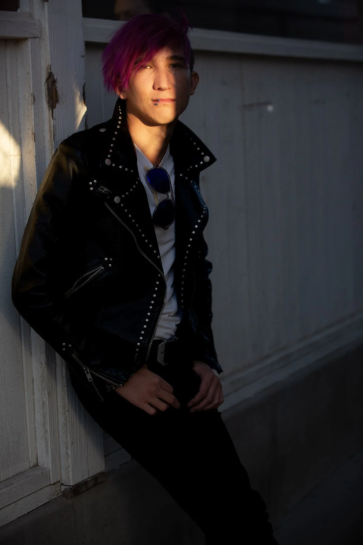 A young person with pink hair leaning against a wooden wall, wearing a black leather jacket with silver studs, white shirt, sunglasses hanging from the shirt, and a lip piercing.
