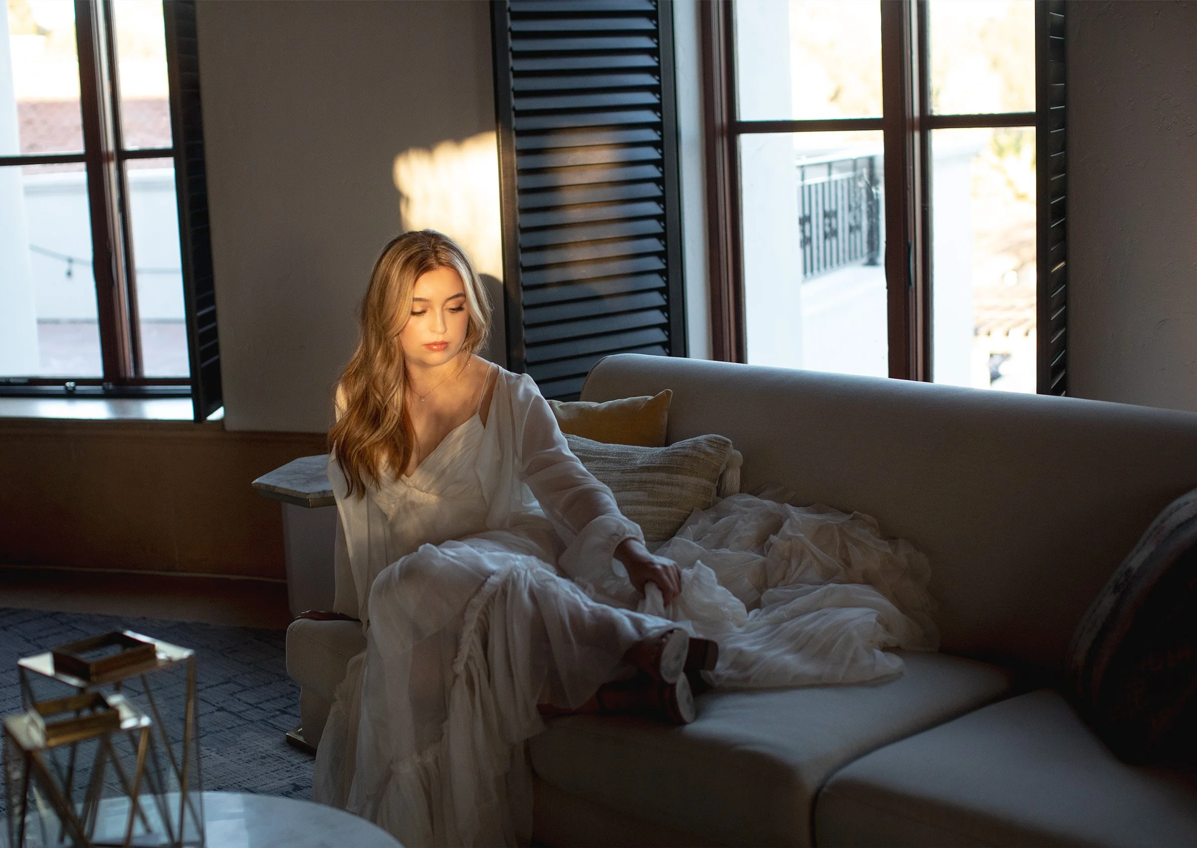 A young woman with long, wavy blonde hair sits on a bed in a room lit by natural sunlight through open windows with black shutters. She is wearing a white blouse and appears to be adjusting her bedding.
