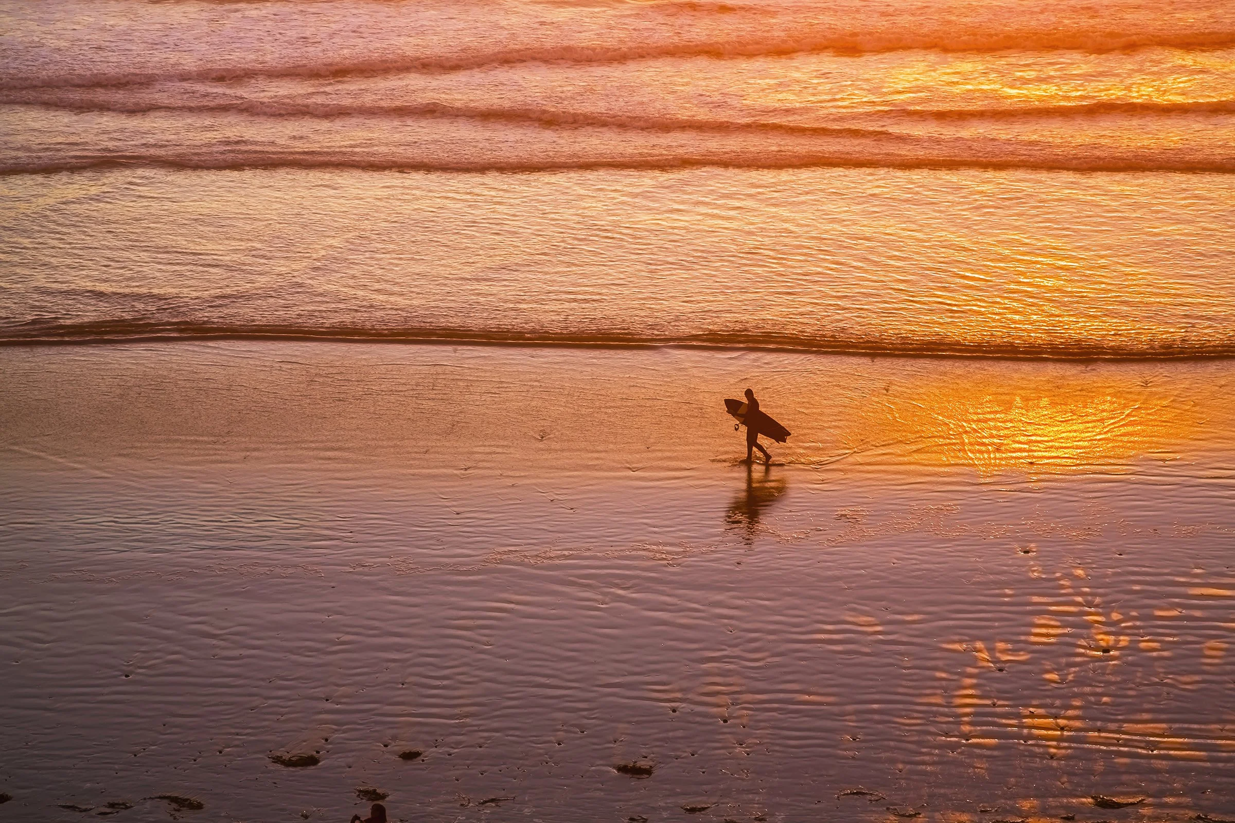 A person carrying a surfboard walking along the shoreline at sunset, with calm waves and a golden-orange sky reflected on the water.