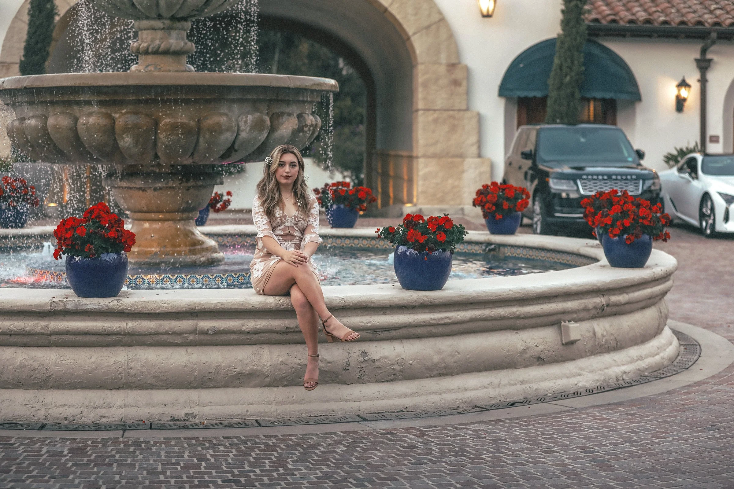 A young woman with blonde hair sitting on the edge of a large decorative fountain with a water feature and red potted flowers. The fountain is in an outdoor courtyard with a building featuring an arched doorway, tiled roof, and parked cars in the background.