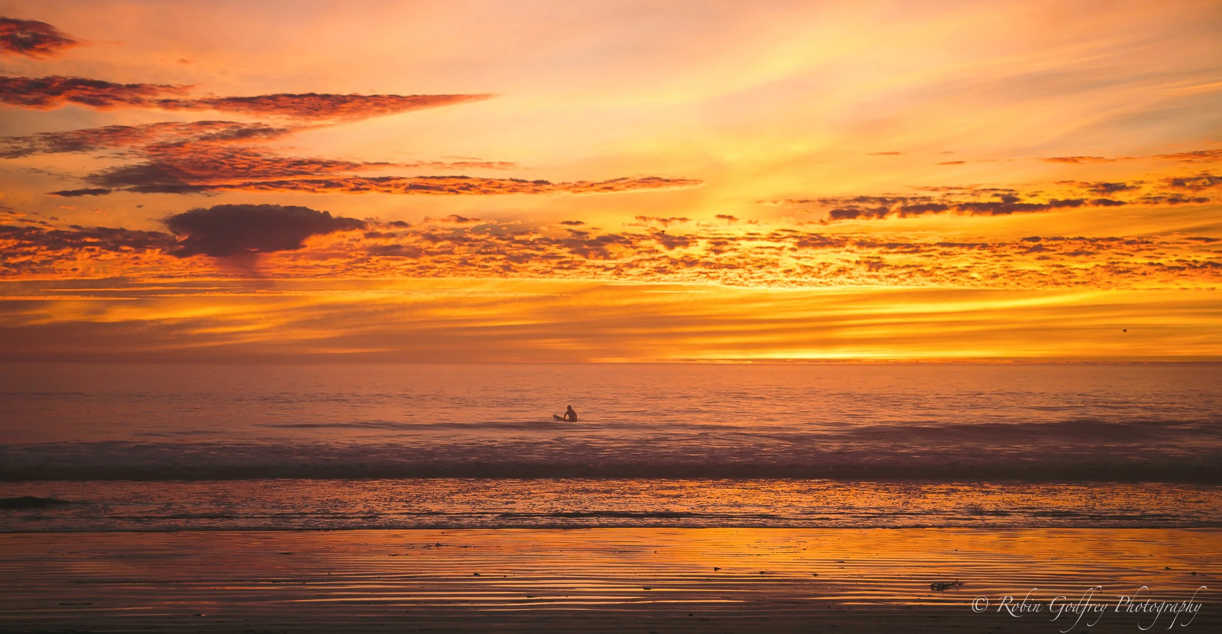 A person kayaking in calm ocean waters during a vibrant sunset with colorful sky and scattered clouds.