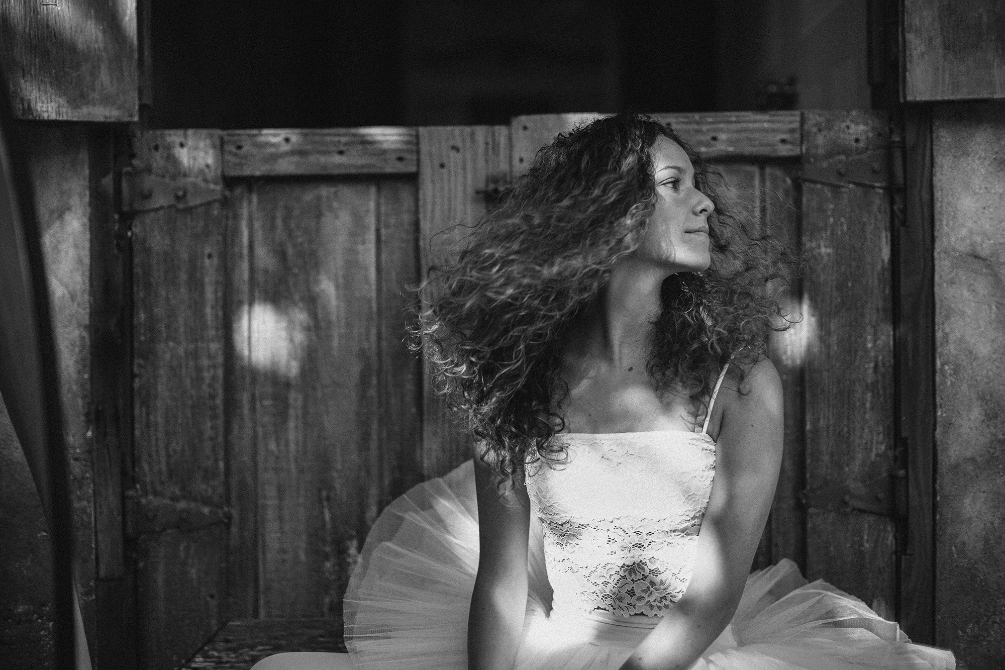 A woman with long curly hair wearing a lace top and tulle skirt, sitting in a wooden enclosure.