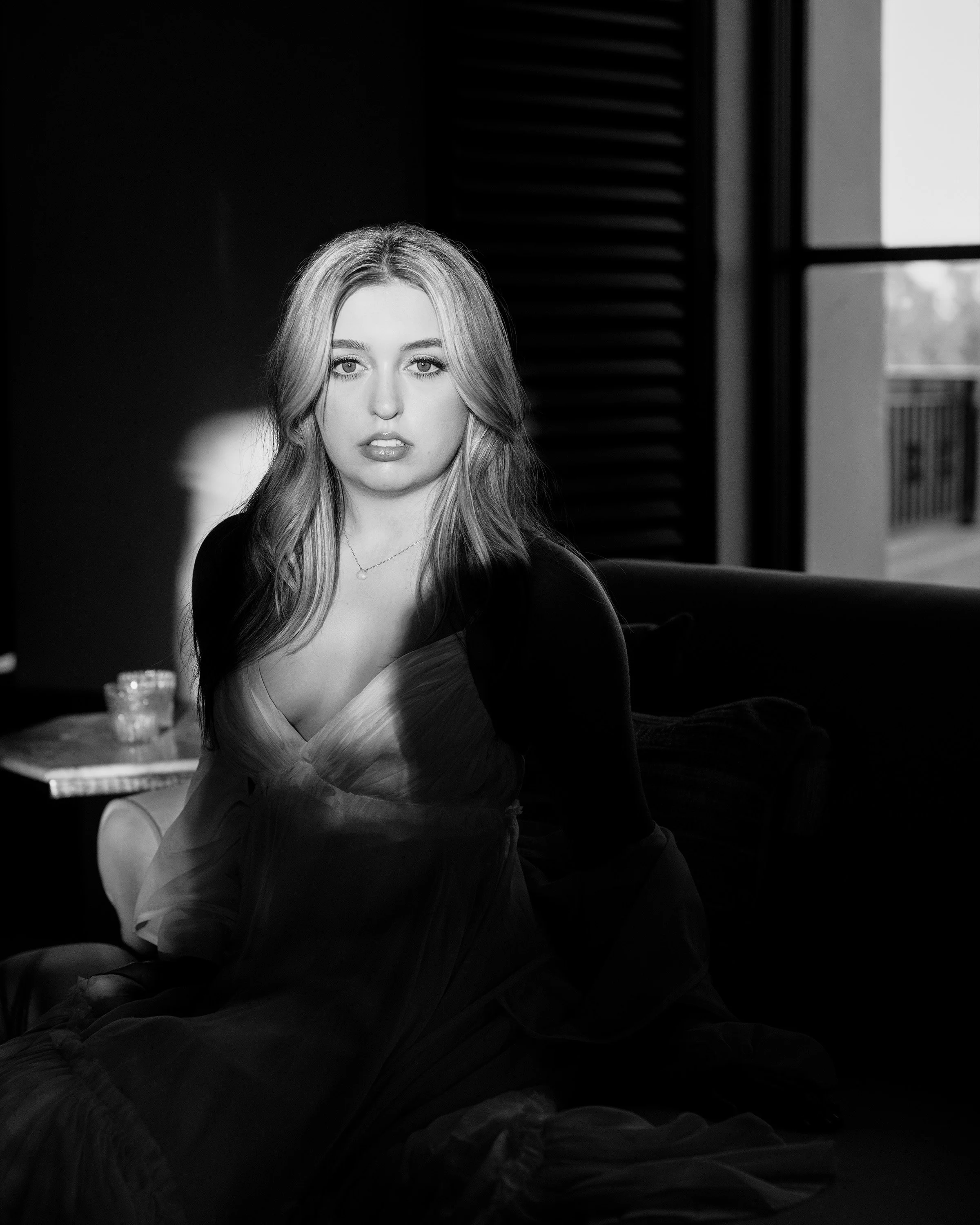 Black-and-white photo of a young woman with long, styled hair, sitting indoors near a window with horizontal blinds. She is wearing a light-colored top and has a neutral expression. Part of a table with a candle and a closed book can be seen in the foreground.