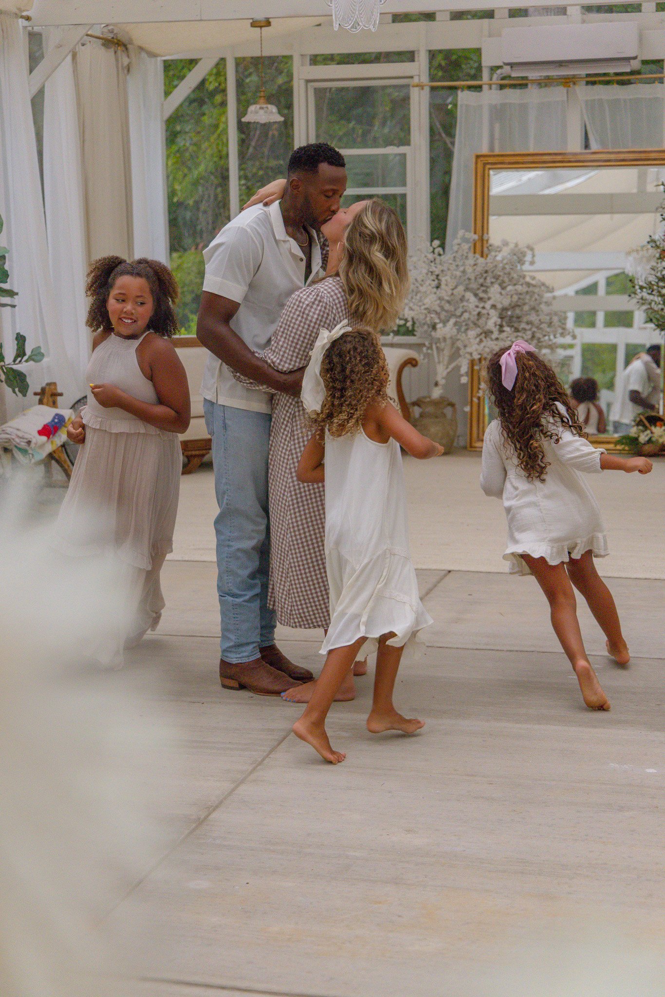 A family of five dancing and kissing in a bright, airy room with large windows and white curtains. The man is kissing the woman, while three young girls dance around them.