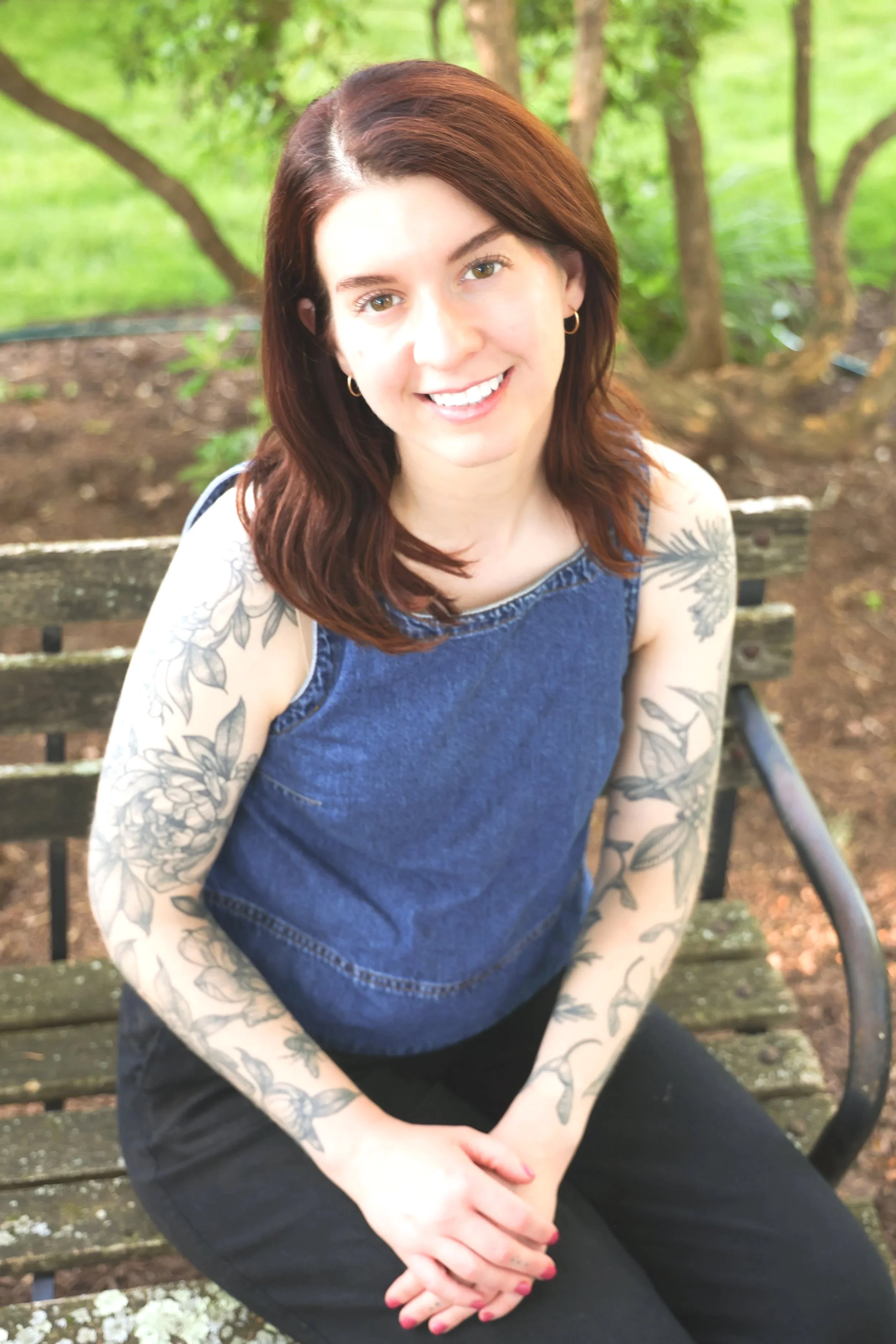 Young woman with red hair and tattoos sitting on a park bench, smiling, with green trees in the background.