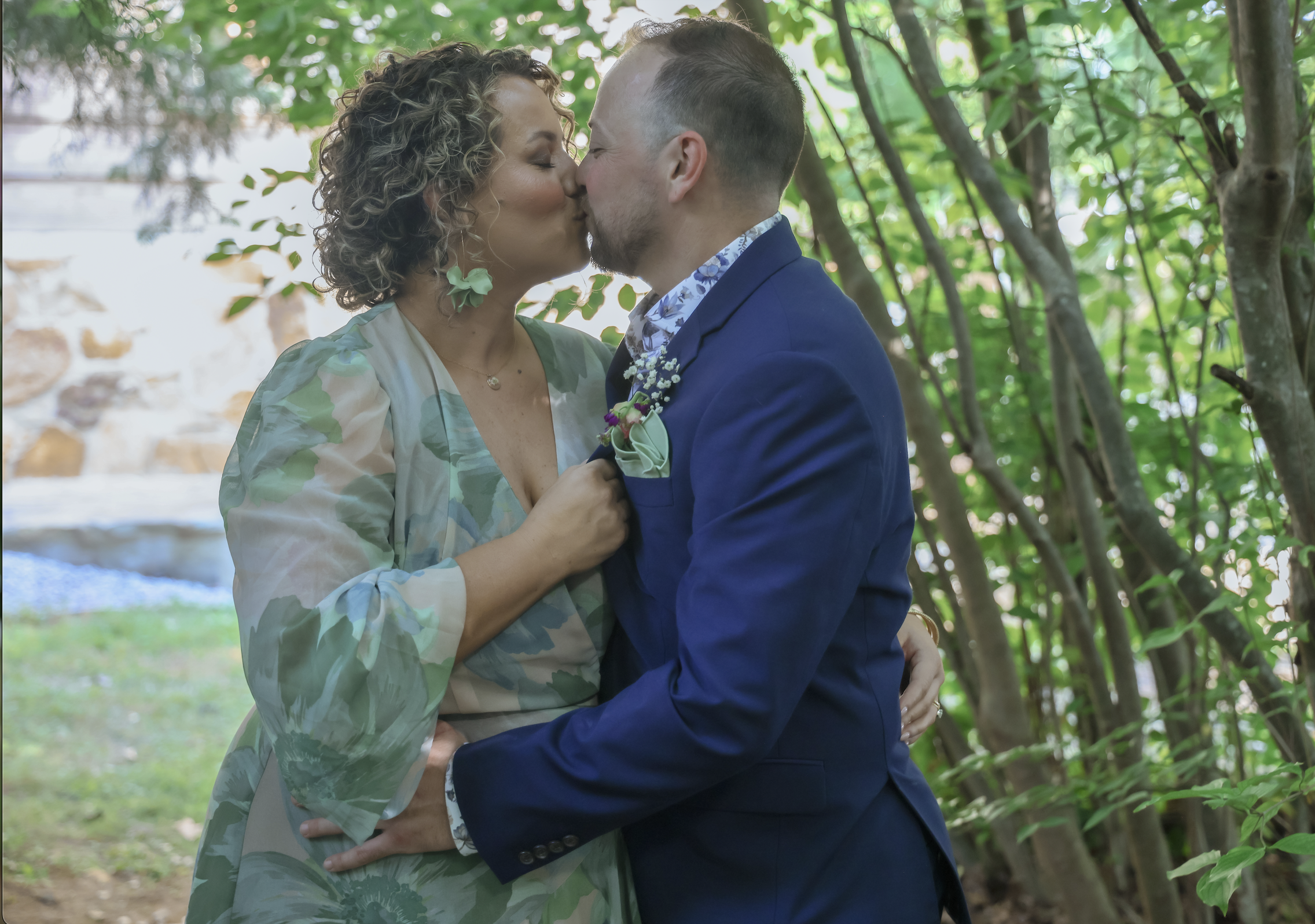 A couple sharing a kiss outdoors among green trees and foliage.
