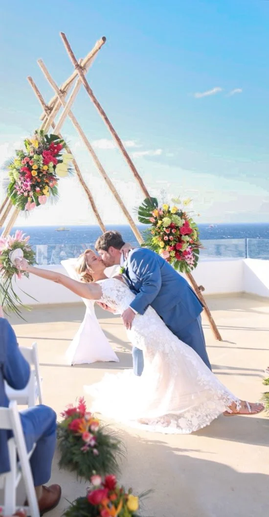 A bride and groom sharing a kiss during their outdoor wedding ceremony on a sunny day, with the ocean in the background and decorated with floral arrangements and a wooden arch.