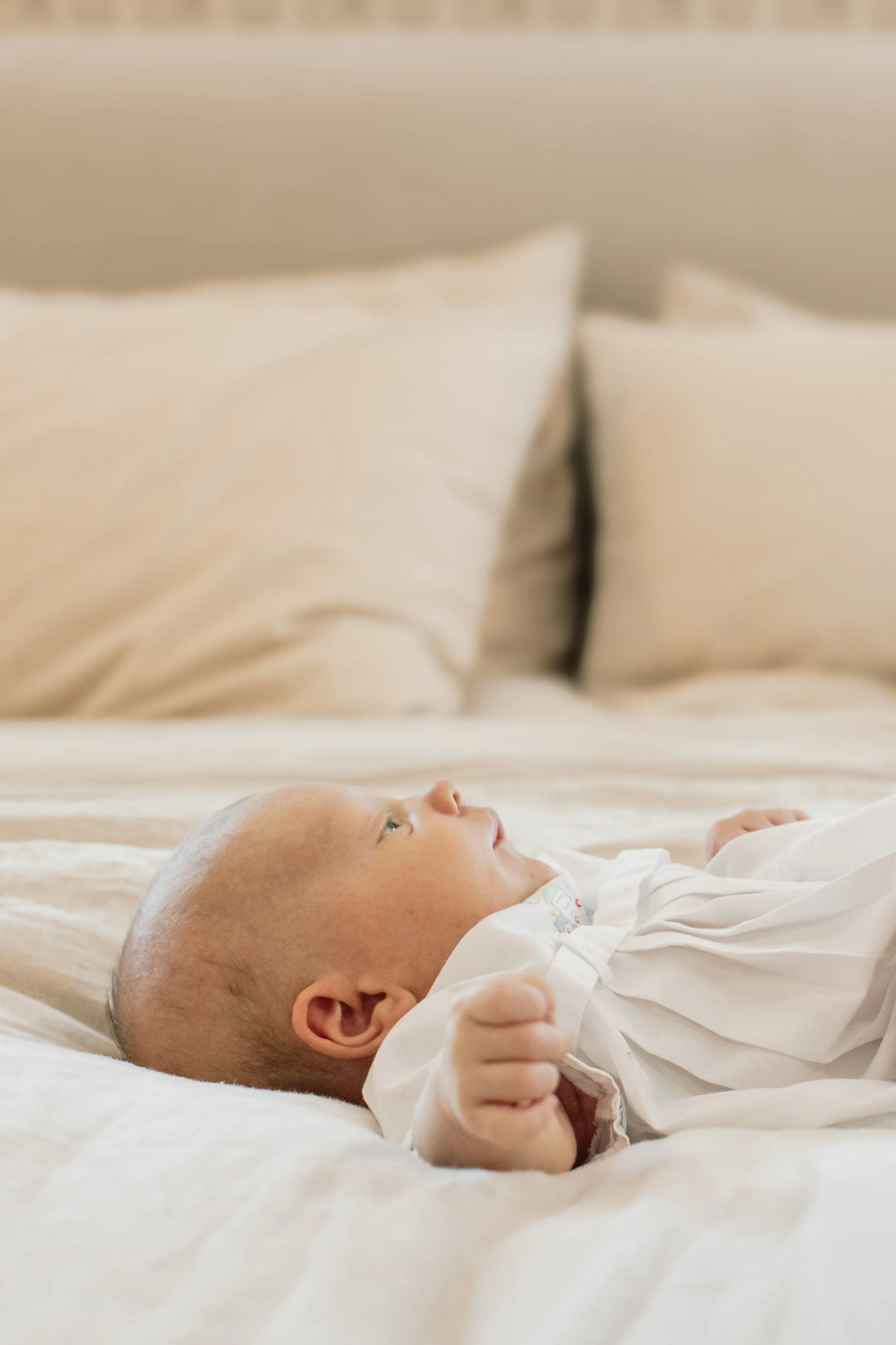 Close-up of a newborn baby laying on a bed with cream-colored pillows in the background, wearing a white outfit with tiny colorful prints, gazing upward.