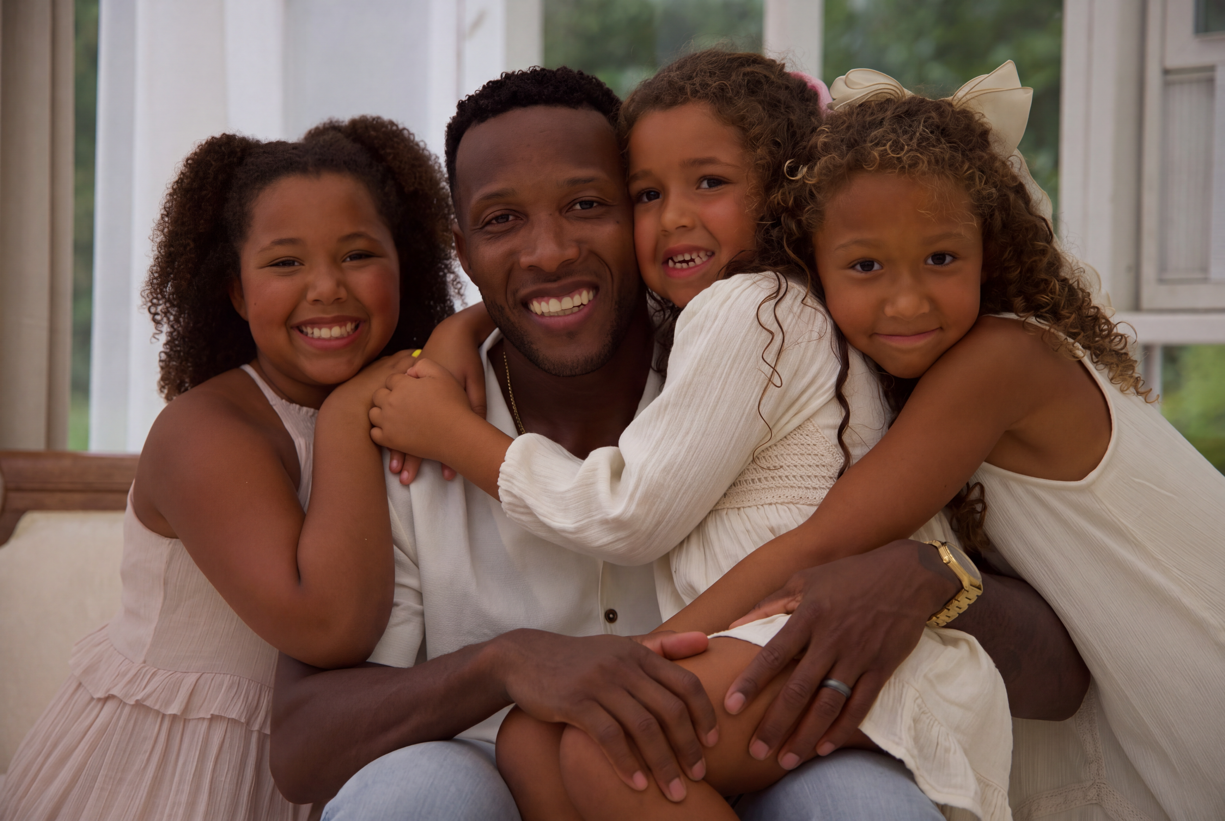 A group of five people, including a man and four young girls, hugging and smiling in a cozy indoor setting with large windows showing greenery outside.