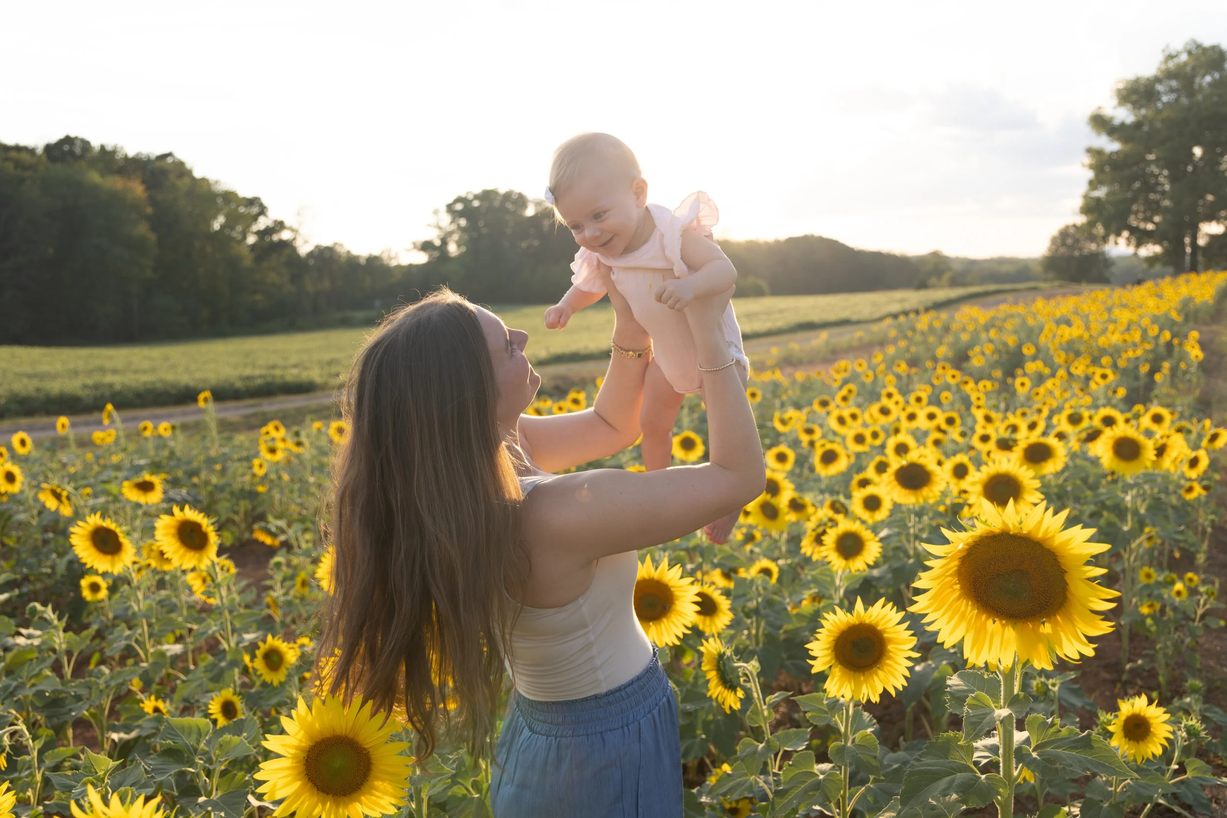 A woman holding a smiling baby in a sunflower field during sunset or sunrise, with trees and open sky in the background.