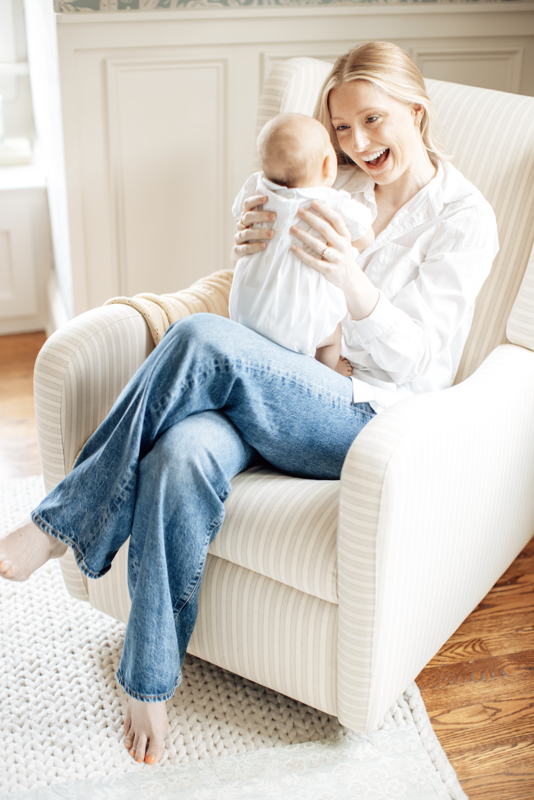 A woman sitting on an armchair, holding a baby and smiling at the baby.