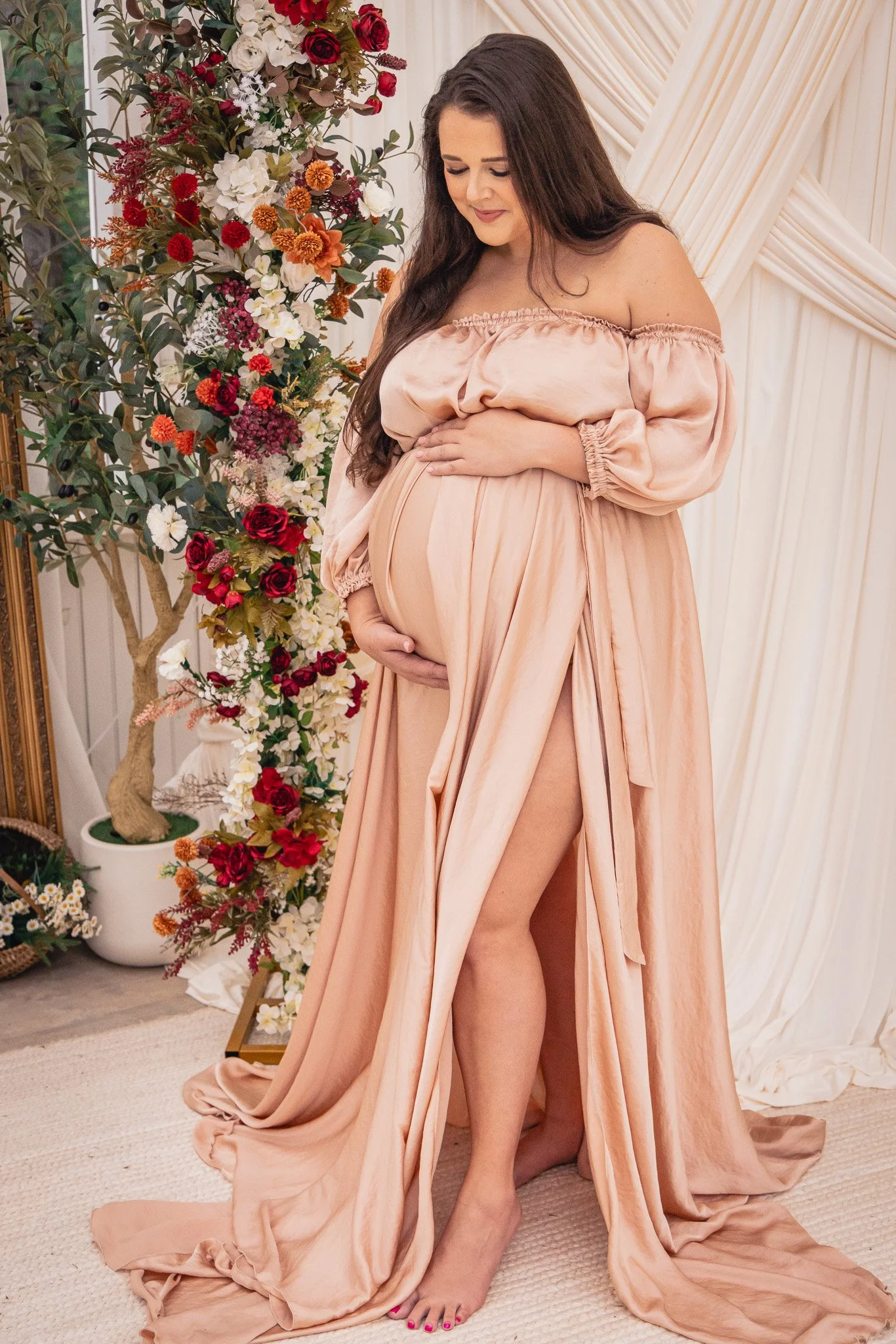 A pregnant woman in a flowing peach-colored dress standing indoors next to a floral arrangement, with her hands on her belly and smiling gently.