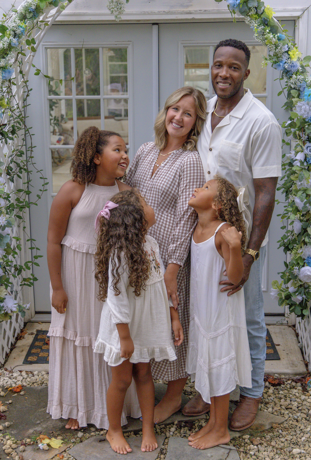 A family gathers under a floral arch outside a house. The group includes a man, a woman, and three young girls, all smiling and looking happy. The setting appears to be a celebratory occasion.