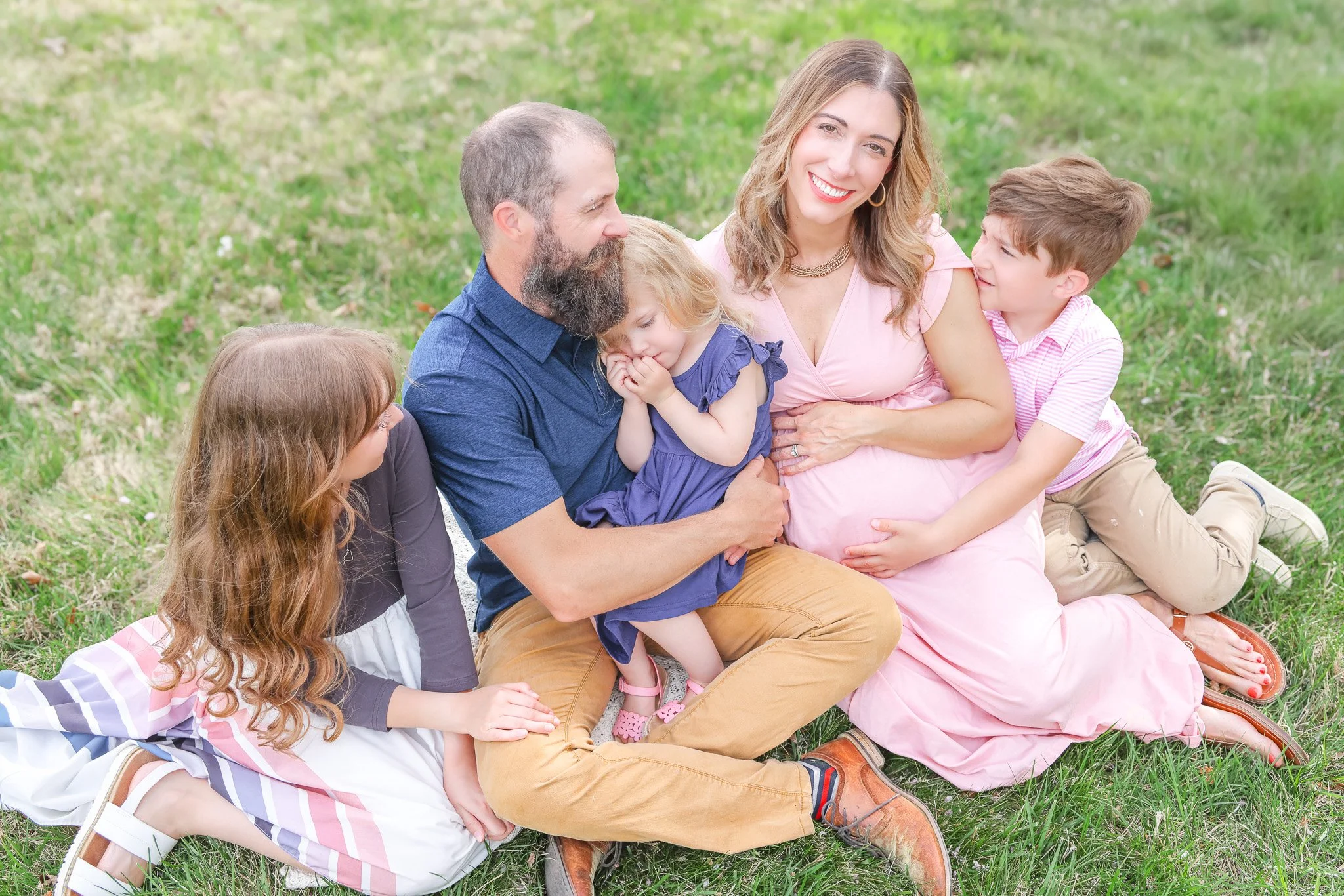 A family of six sitting on grass, showing a pregnant woman, a man with a beard, and four children, smiling and enjoying outdoors.