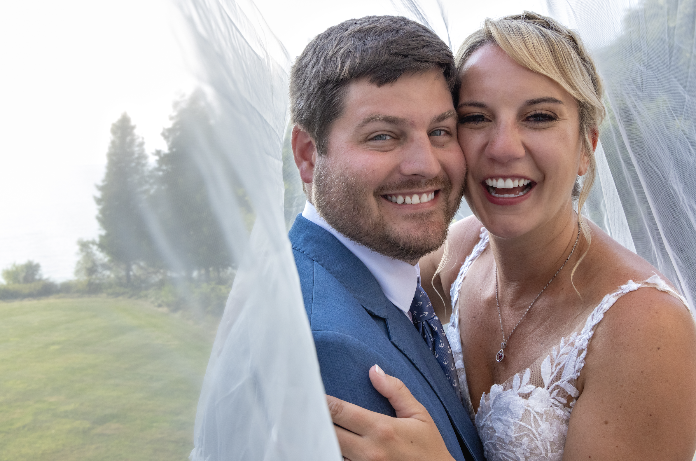 A happy couple, a man in a blue suit and a woman in a white lace wedding dress, posing closely together outdoors, smiling at the camera.