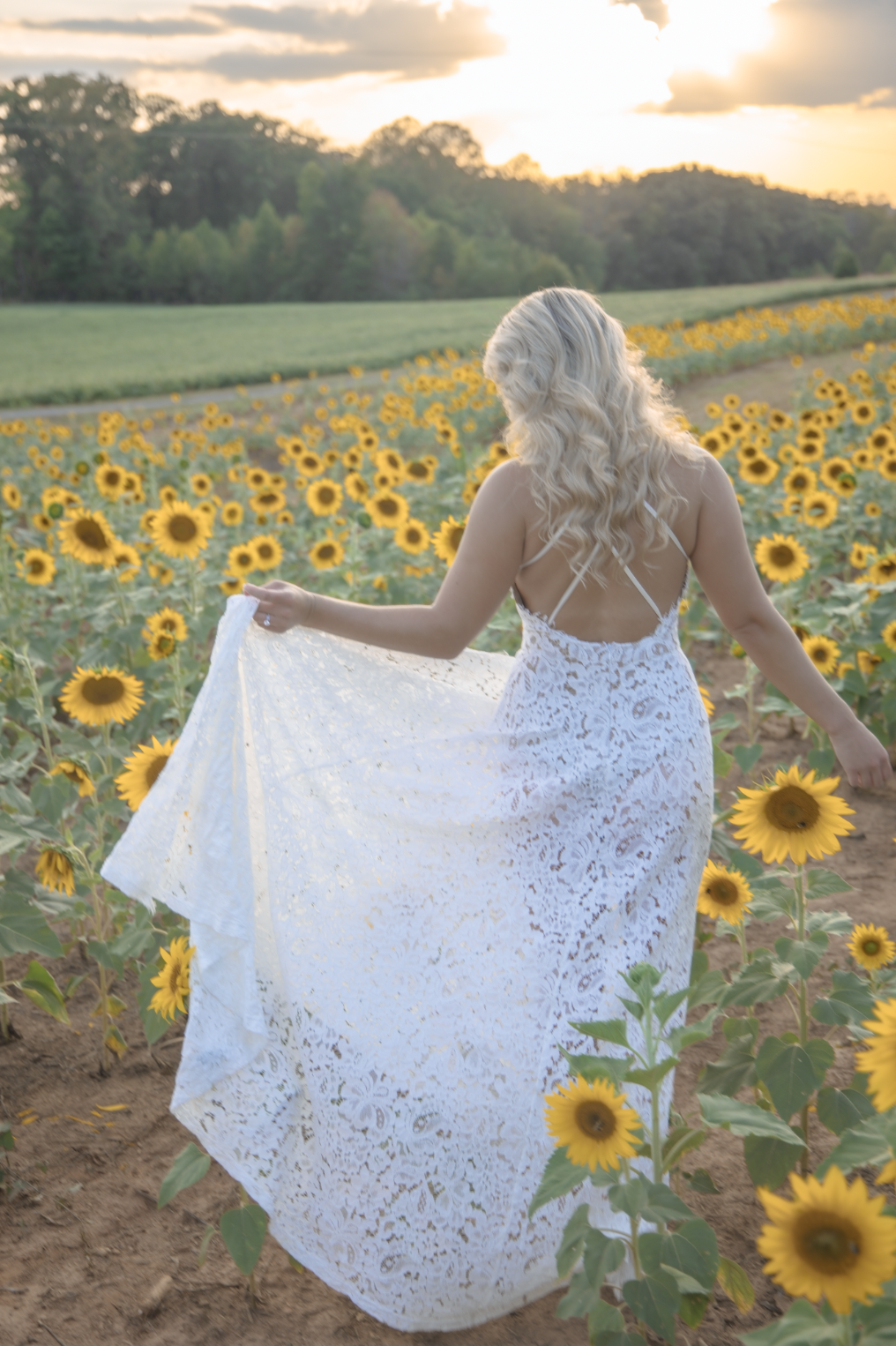 A woman in a white lace dress walking through a sunflower field at sunset with a scenic background of trees and a cloudy sky.