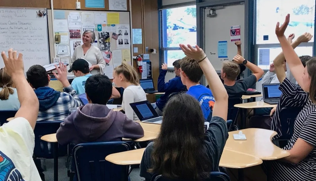 Classroom scene with students raising hands, a teacher smiling at the front, whiteboard, and educational posters on the walls.