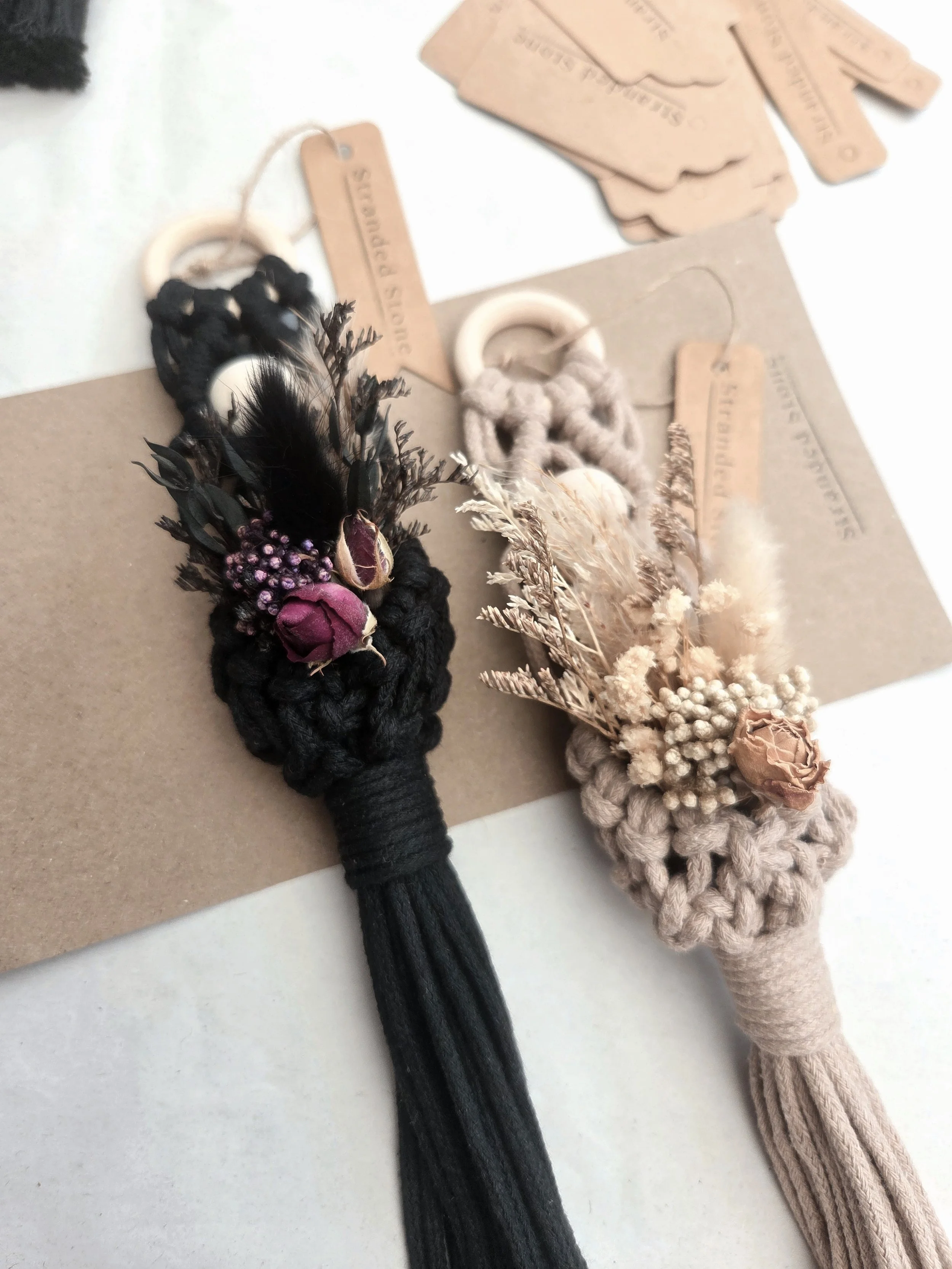 Two knotted hair accessories, one black with dried flowers and shells, the other beige with dried flowers, displayed on a tan and white background with tags reading shuttle puncture.