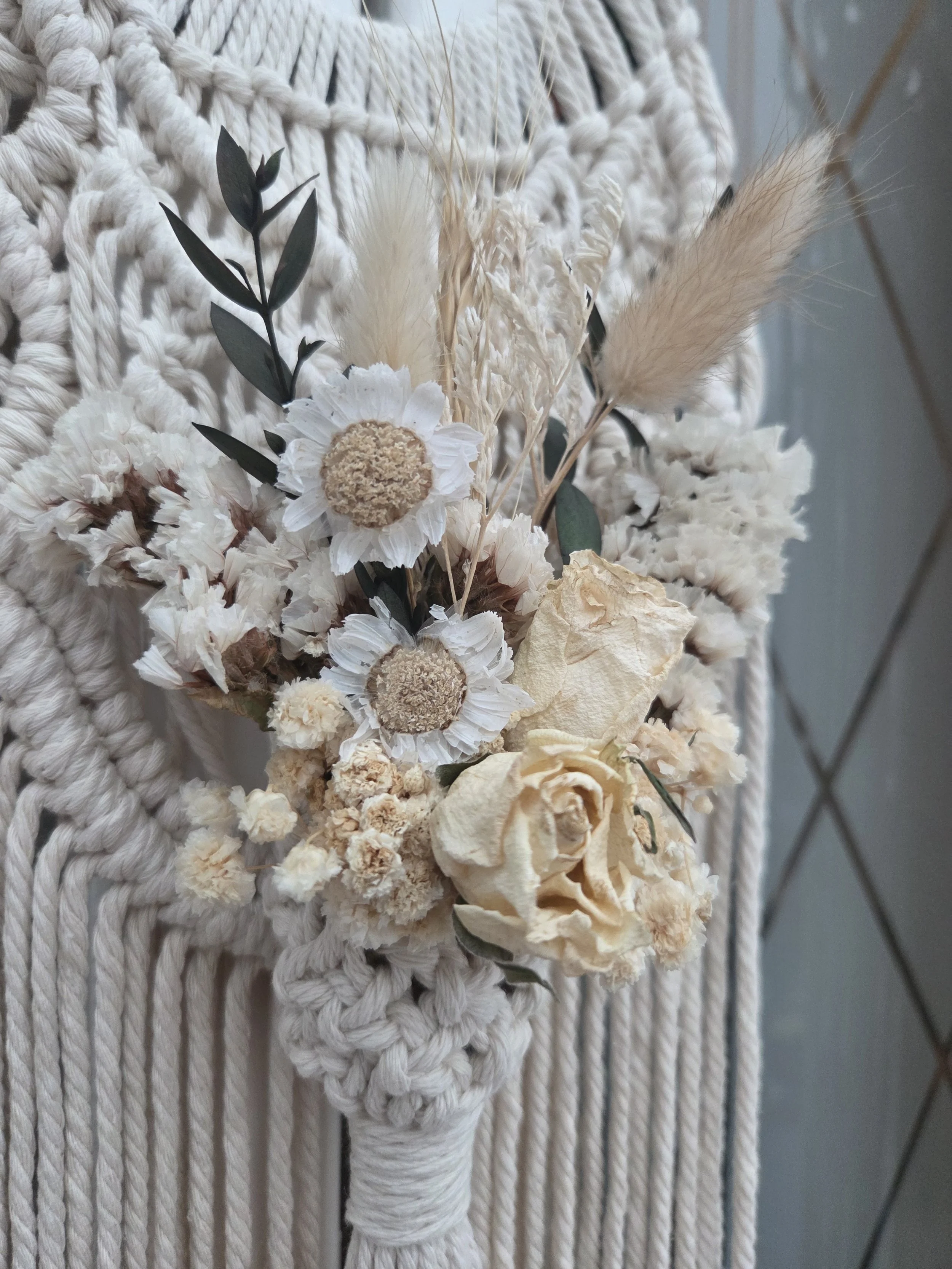 A bouquet of dried white flowers with green leaves and beige pampas grass, wrapped with white rope, hanging on a white woven textured wall.