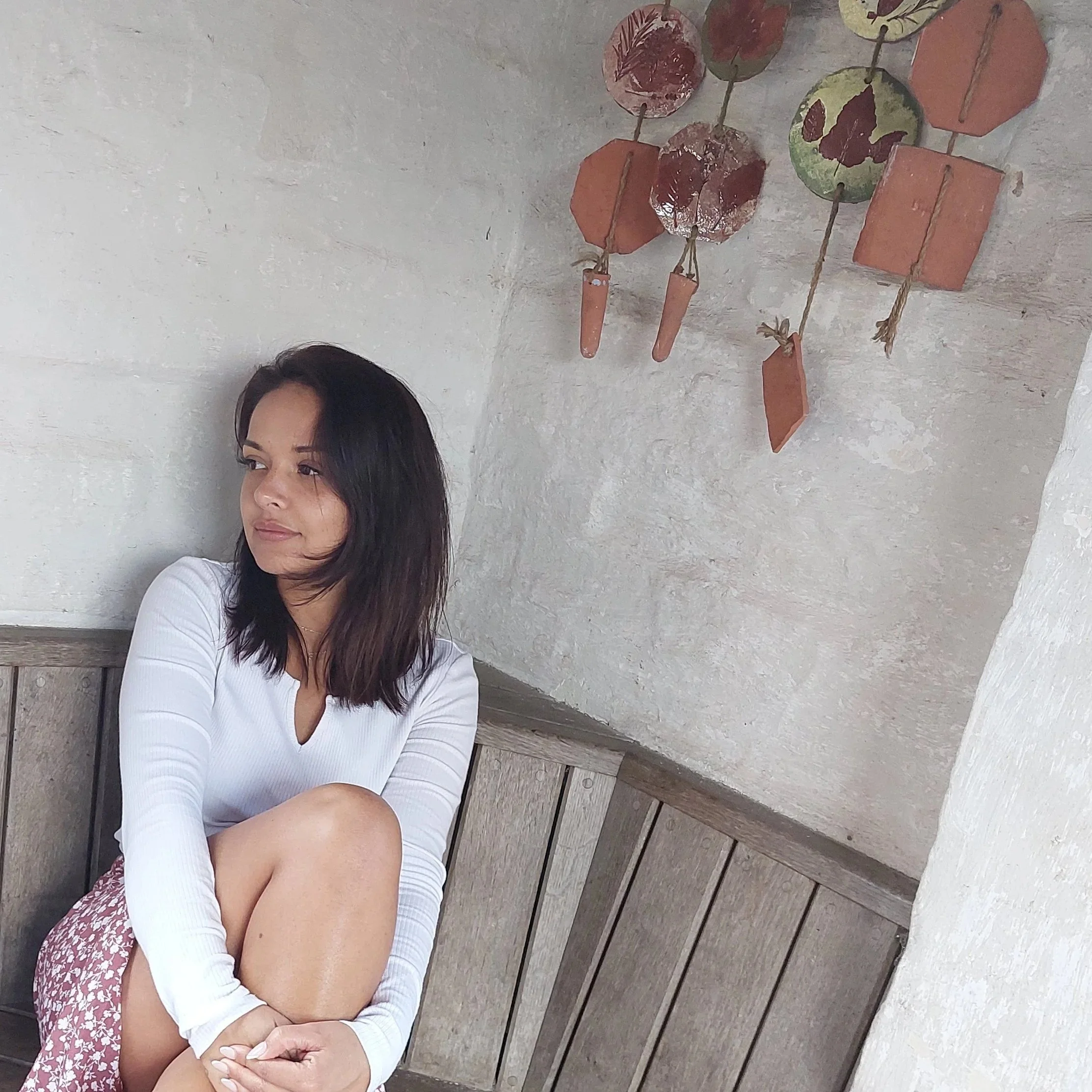 A woman with black hair, wearing a white long-sleeve top, sitting on a wooden bench with a white wall behind her. Part of her face and body is visible.