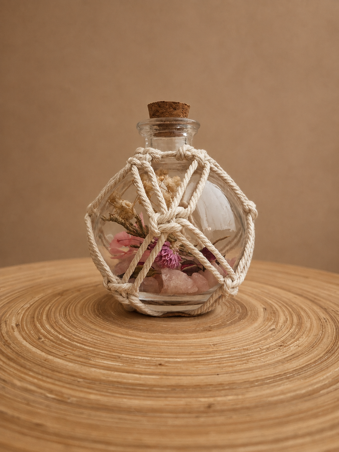 A glass bottle with a cork stopper, decorated with a white rope net, and containing pink flowers and crystals, placed on a wooden surface.