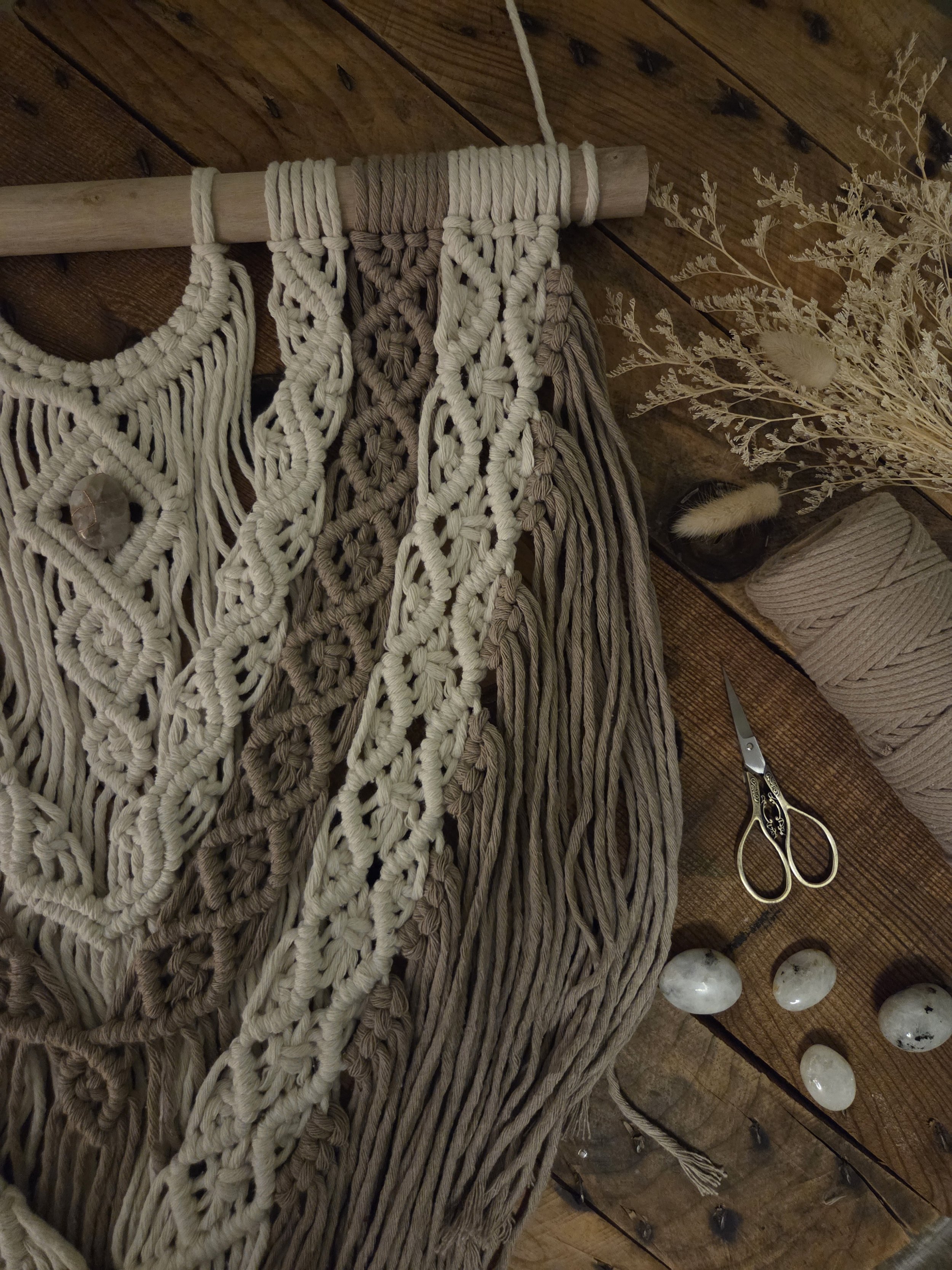 A macramé wall hanging in cream and beige hanging on a wooden pole, with a set of scissors, a bundle of dried flowers, and some polished stones on a wooden surface nearby.