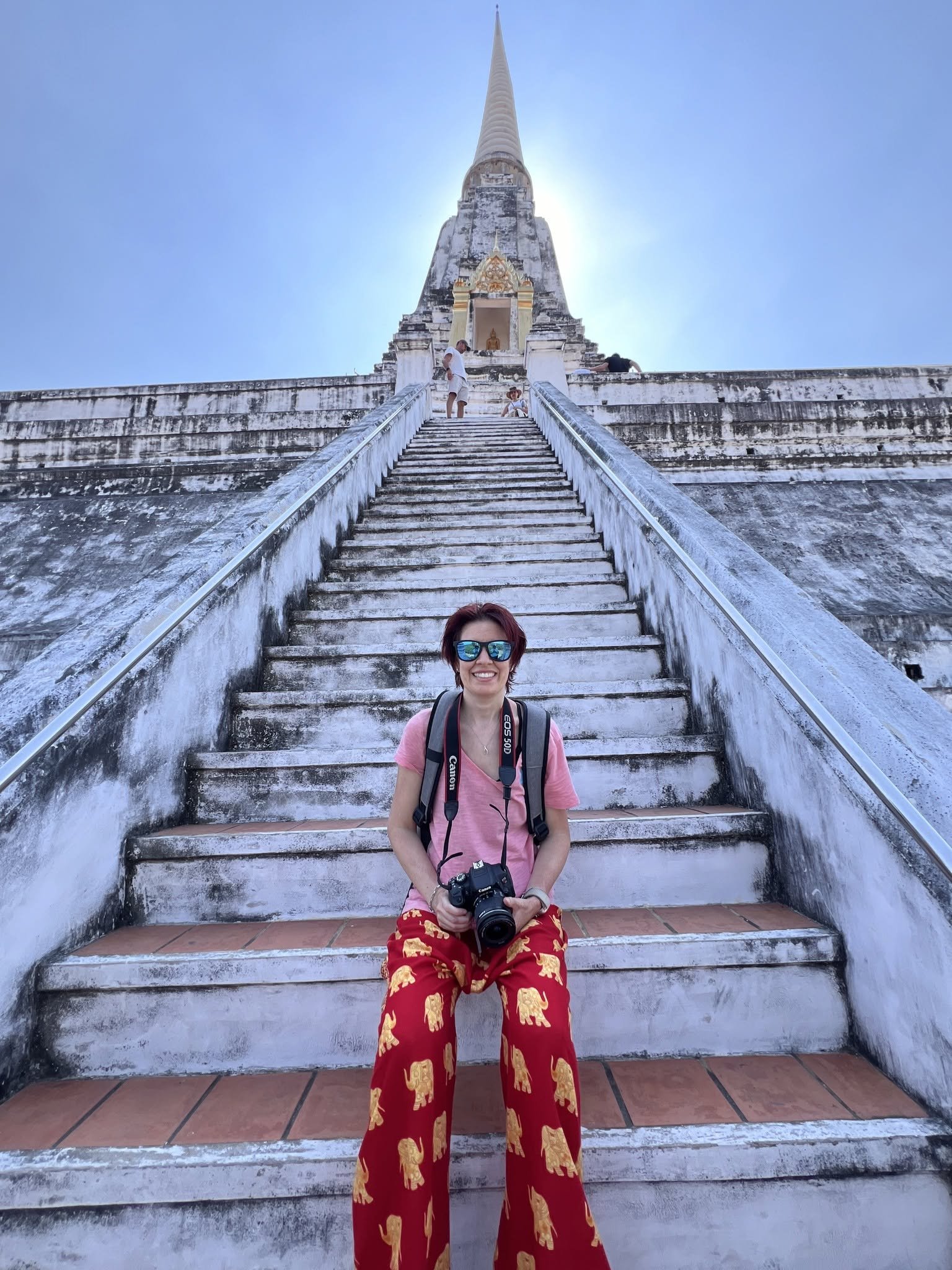 Woman sitting on stone steps leading up to a white temple with a tall spire, holding a camera, wearing red pants with yellow elephants, sunglasses, and a pink shirt, with other visitors nearby and a bright blue sky.