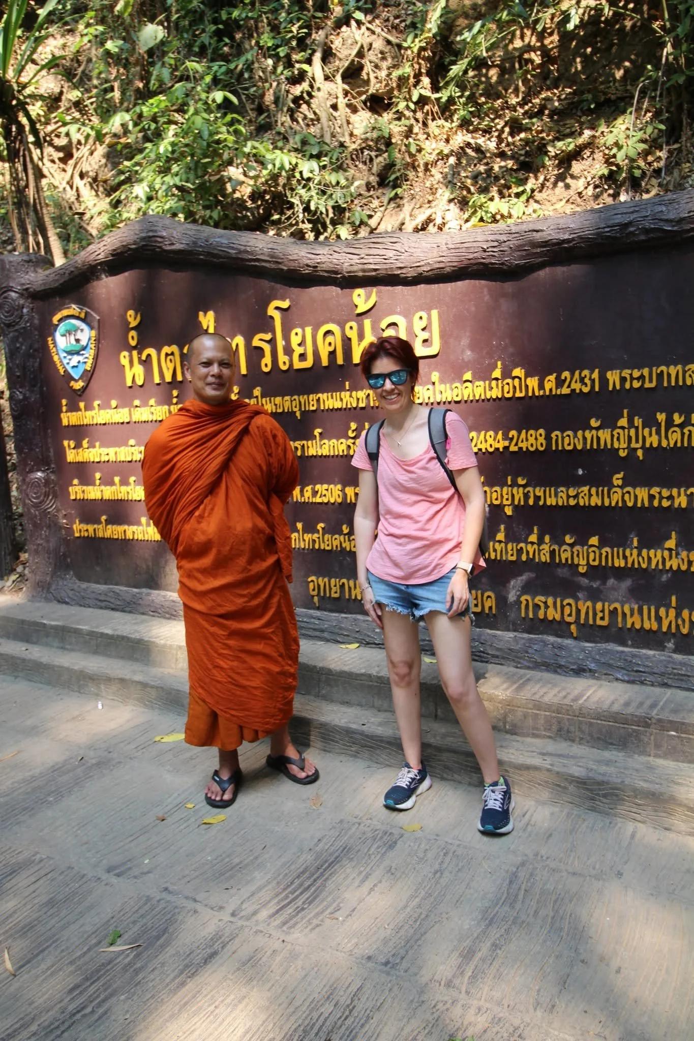 A woman in sunglasses and casual clothing standing next to a Buddhist monk in orange robes in front of a large wooden sign with Thai writing. The sign has a crest logo and is situated outdoors with greenery in the background.