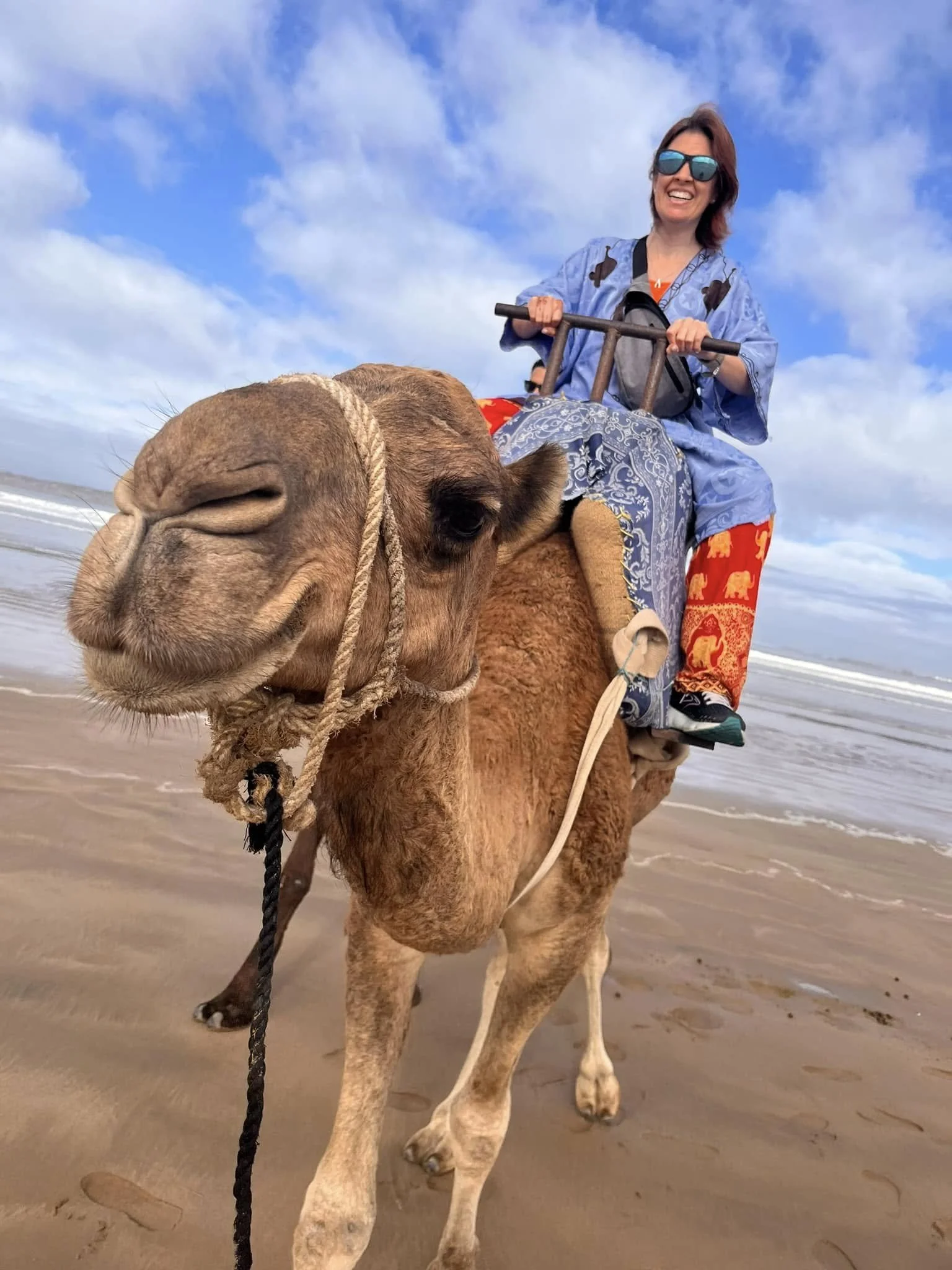 A woman riding a camel on a beach, smiling and wearing sunglasses, with the ocean and cloudy sky in the background.