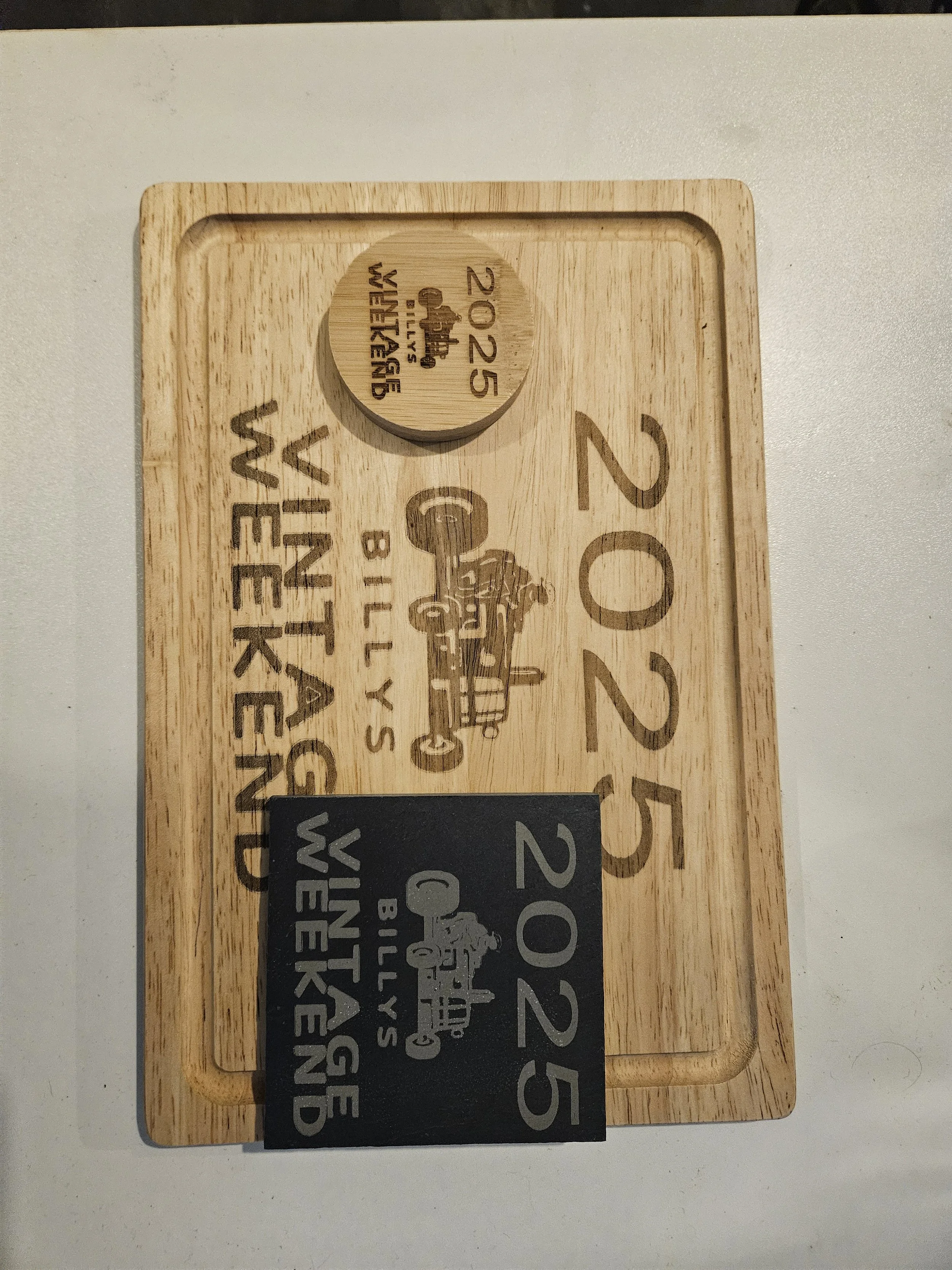 A wooden tray holding two coasters with engraved designs, one exposing part of a chalkboard-style sign with text.