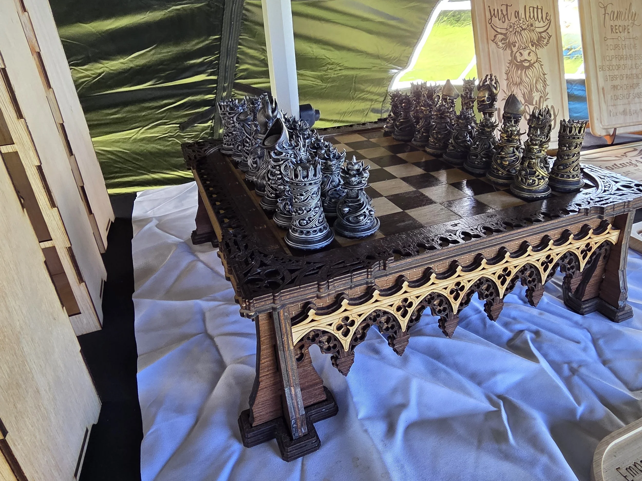 A wooden chess set with intricately carved black and white pieces on a decorative chessboard. The setting appears to be outdoors or in a tent, with a painted wooden sign in the background that reads 'Just a little family recipe' and includes a list of ingredients.