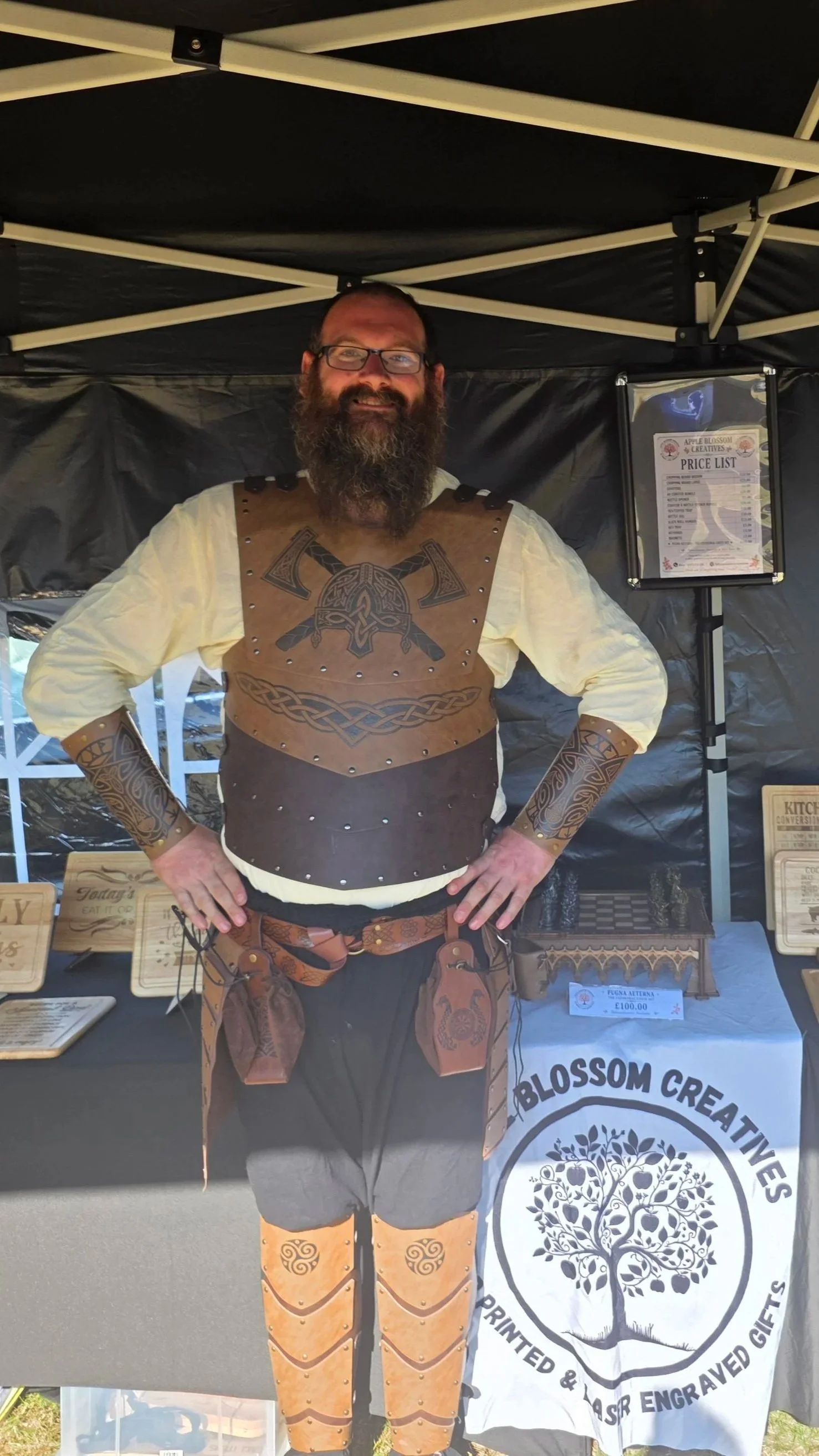A man with a beard and glasses standing behind a table at a craft fair, dressed in medieval-style leather armor with intricate designs, under a black canopy. There are signs and items displayed on the table.