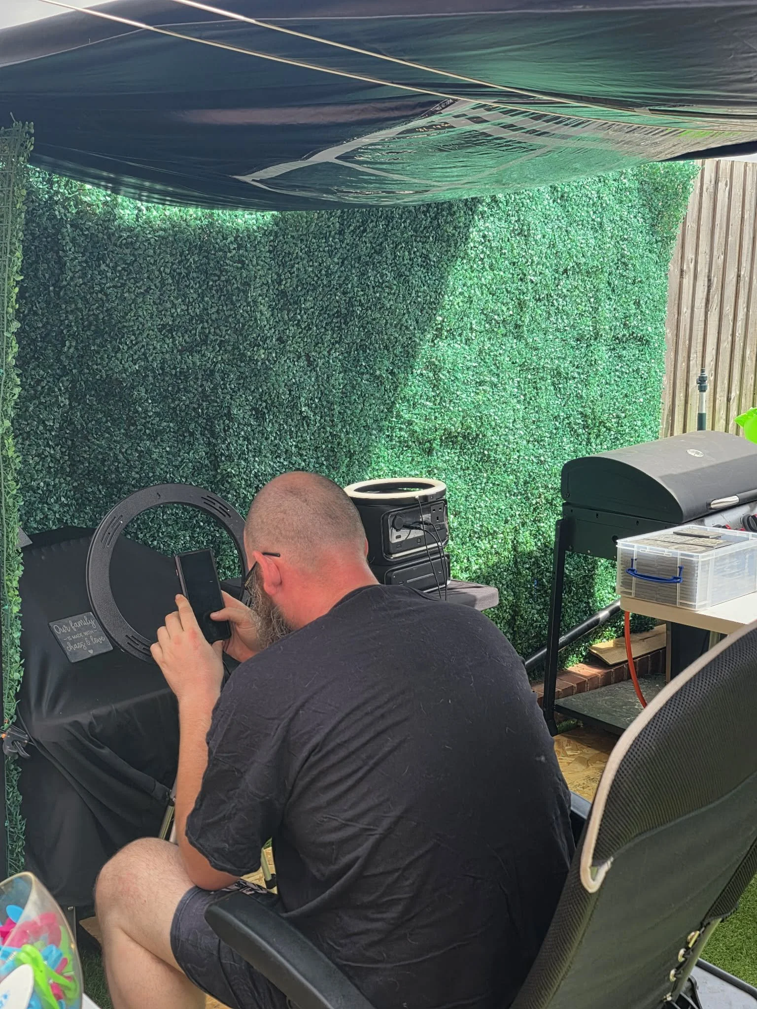 A man with a beard and glasses sitting outside, looking at a smartphone, with a green artificial hedge and wooden fence behind him, and a speaker and plastic storage container on a table nearby.