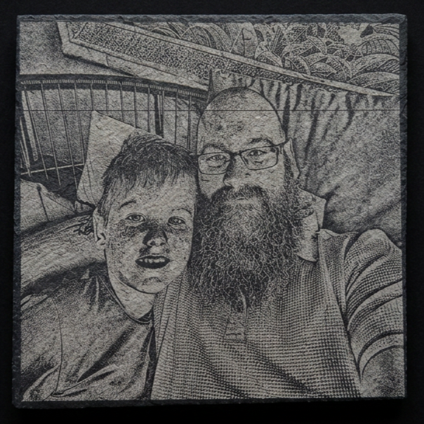 A black and white photo of a man with glasses and a beard taking a selfie with a young boy, both lying on beds with patterned sheets.