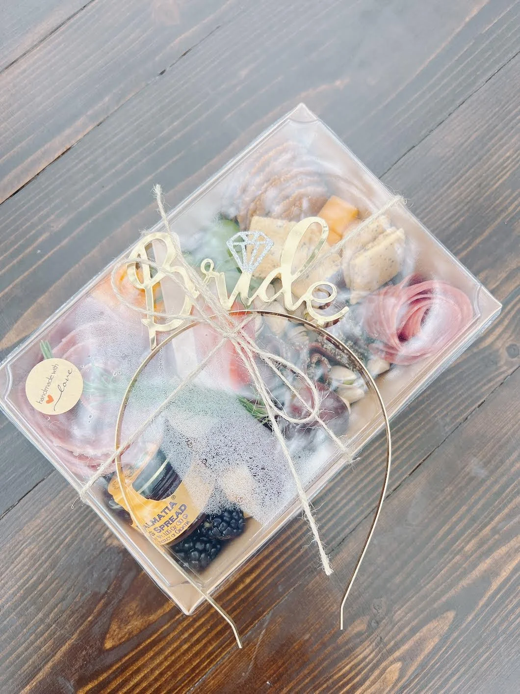 Clear gift box containing an assortment of snacks and cheese, decorated with a gold 'Bride' sign and a small stick with a note that says 'Handmade with Love.' The box is tied with twine and placed on a wooden surface.