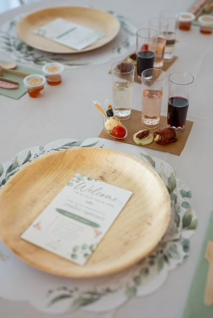 Table setting with a wooden plate, a menu, and a tasting flight of nine small glasses of various colored liquids with some small food items. Honeyberry charcuterie workshop
