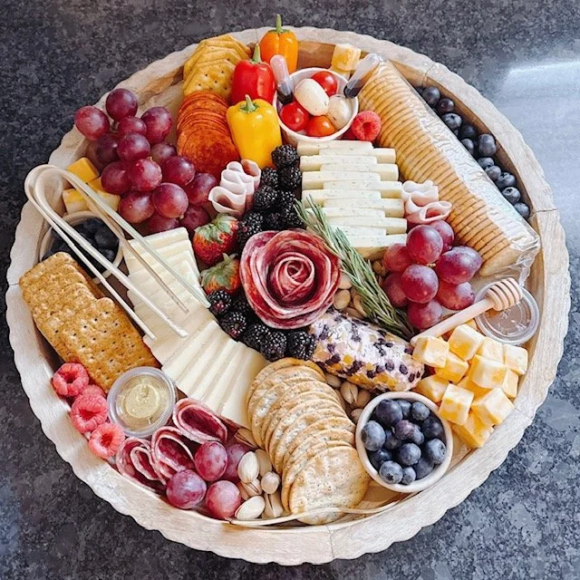 A circular cheese and fruit platter featuring grapes, blueberries, strawberries, blackberries, sliced cheese, cheese blocks, crackers, sliced meats, candies, and a decorative edible flower with rosemary.