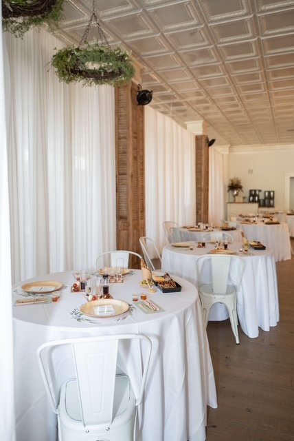 Round tables with white tablecloths set with plates, glasses, and cutlery in a decorated event space with curtains, hanging greenery, and wood-paneled walls.