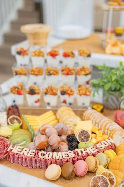 Dessert table with mini cheesecakes topped with fruit, fresh fruit, fruit slices, macarons, cookies, and a decorative 'HERE COMES THE SEASON' sign.