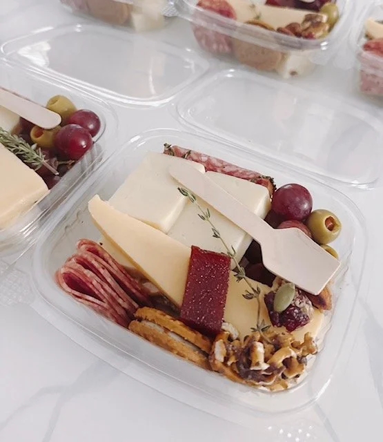 Plastic food container with assorted cheeses, grapes, sliced meat, nuts, and a small wooden cheese knife.