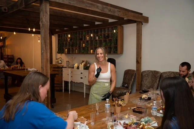 A woman with blonde hair, wearing a white top and green skirt, smiling and speaking to a group at a dinner party in a cozy, rustic indoor setting.