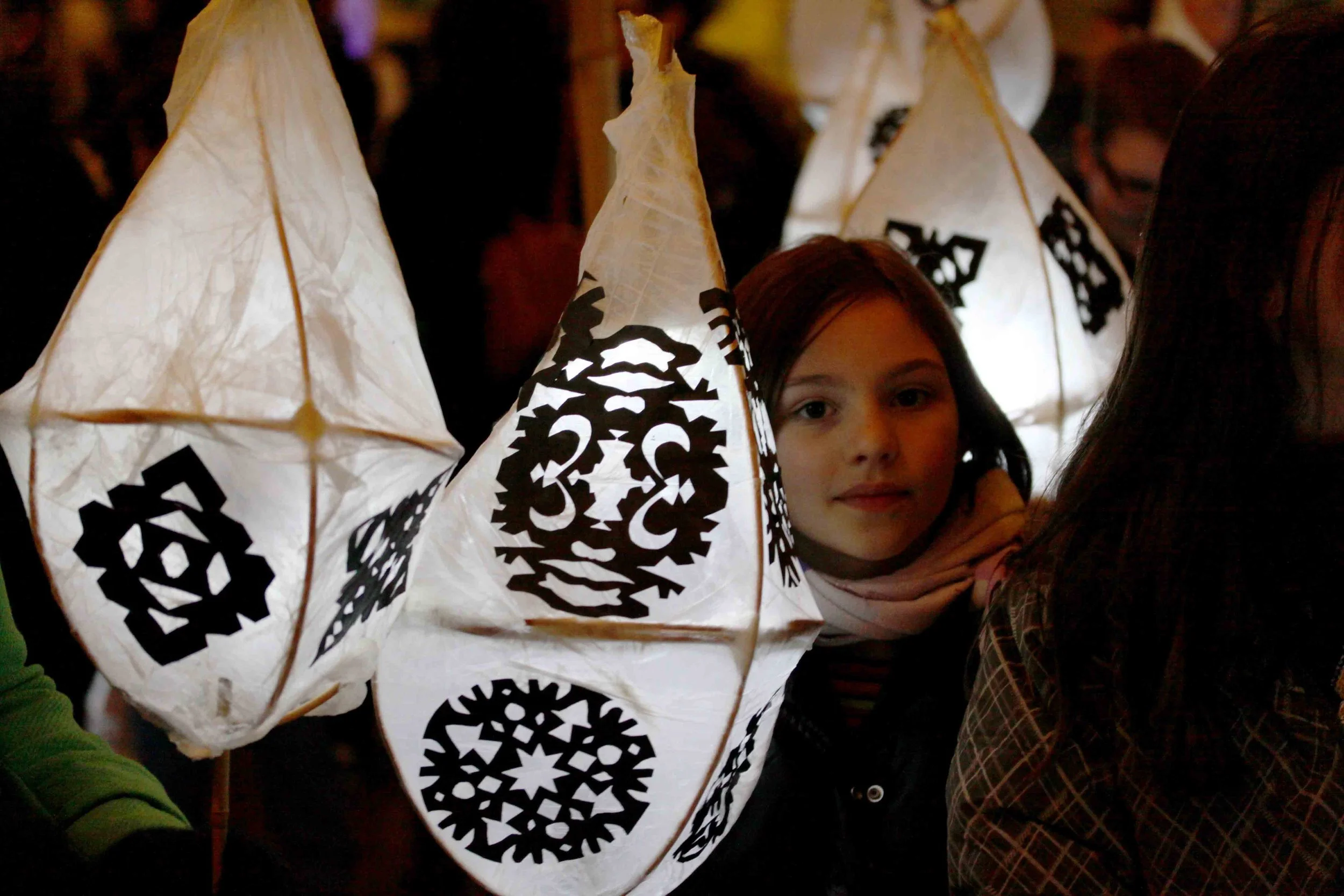 A young girl with brown hair and brown eyes is partially hidden behind a white lantern with black decorative patterns. The lantern is illuminated from inside and surrounded by other similar lanterns, likely during a nighttime festival or celebration.