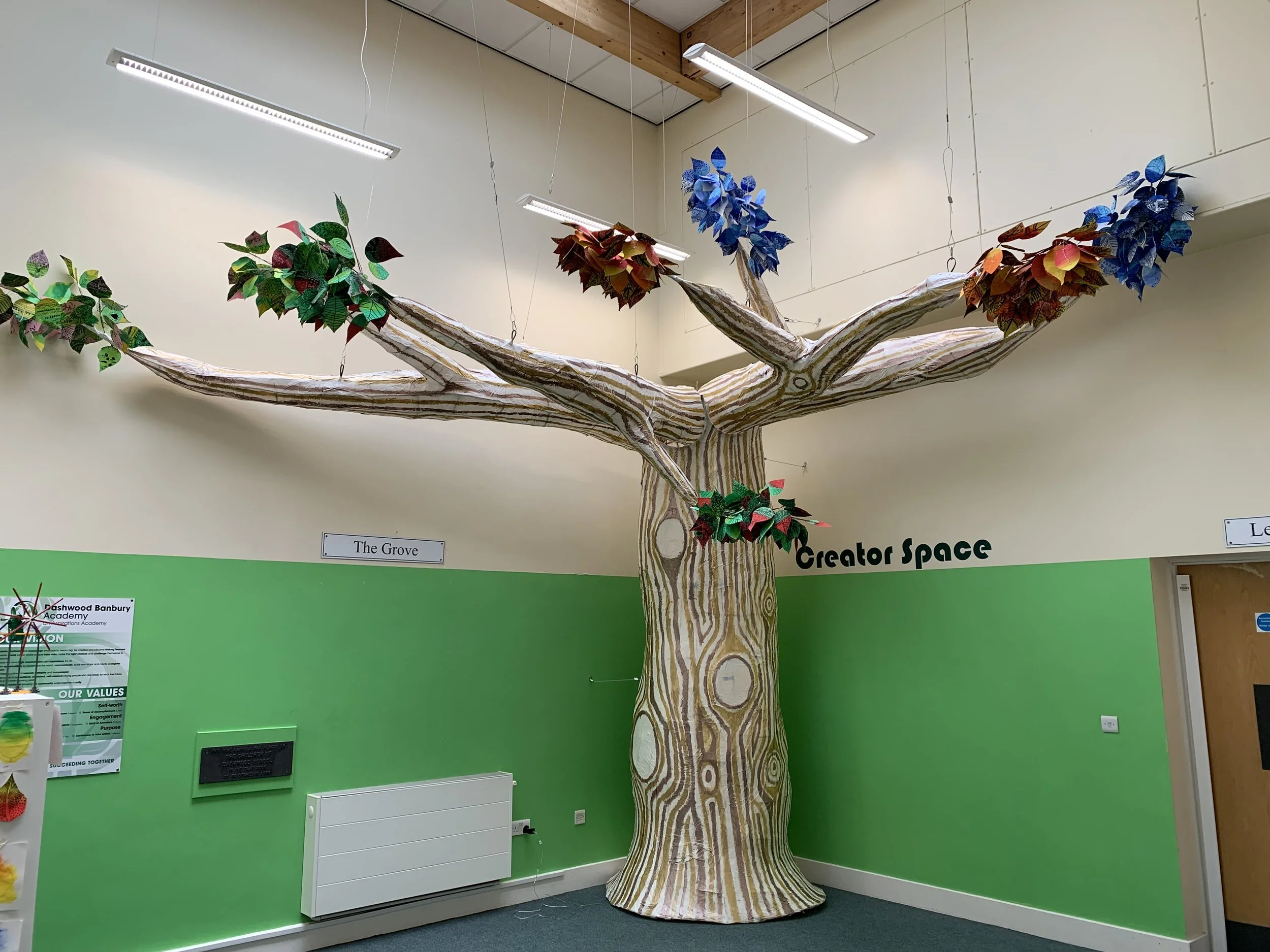 An indoor display of a large artificial tree with a striped trunk and colorful paper leaves hanging from its branches. The background wall is painted green with various informational posters and a white cabinet visible.