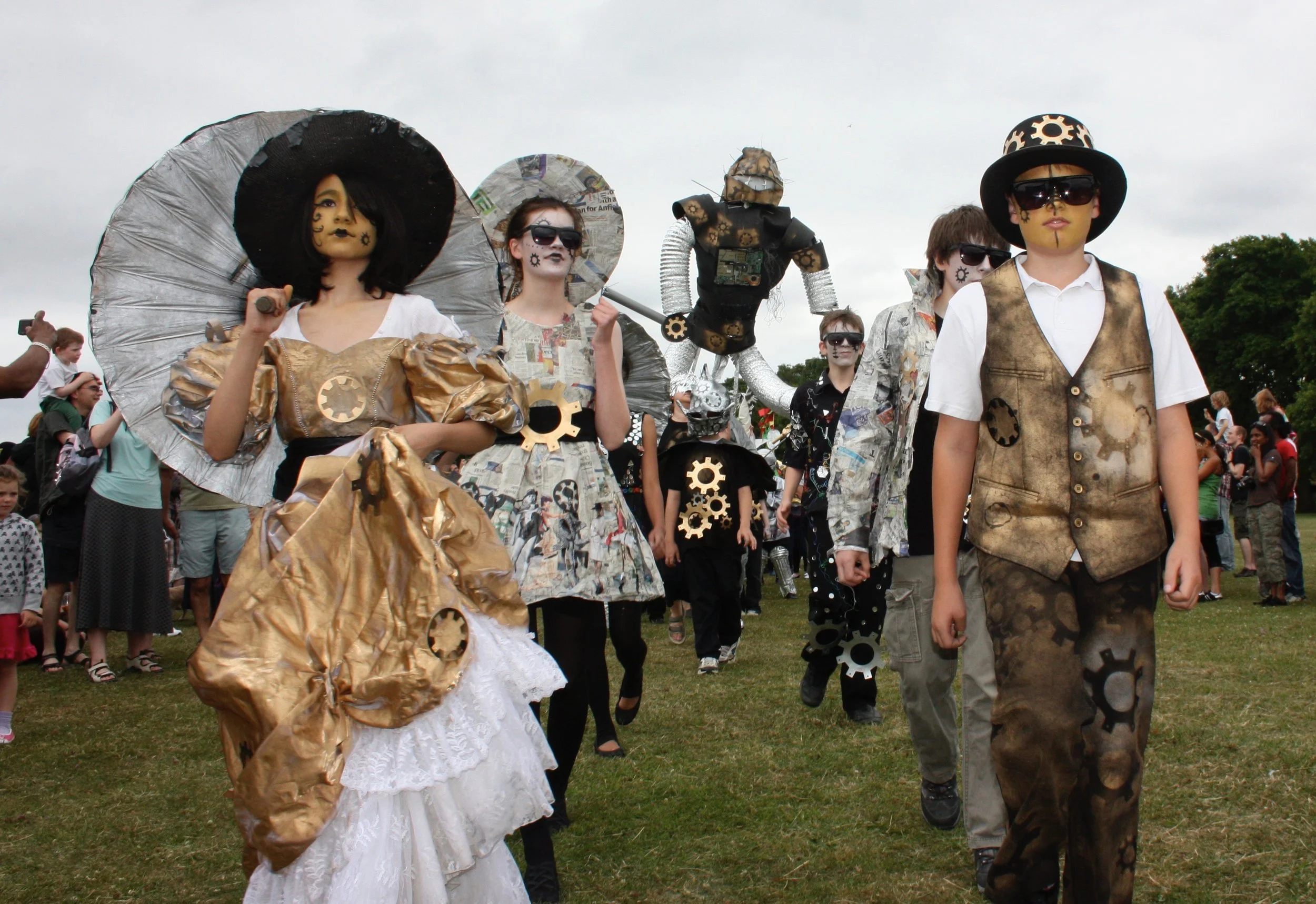 People dressed in steampunk costumes with gears, newspaper print, and metallic accessories walking outdoors on a cloudy day, with onlookers in the background.